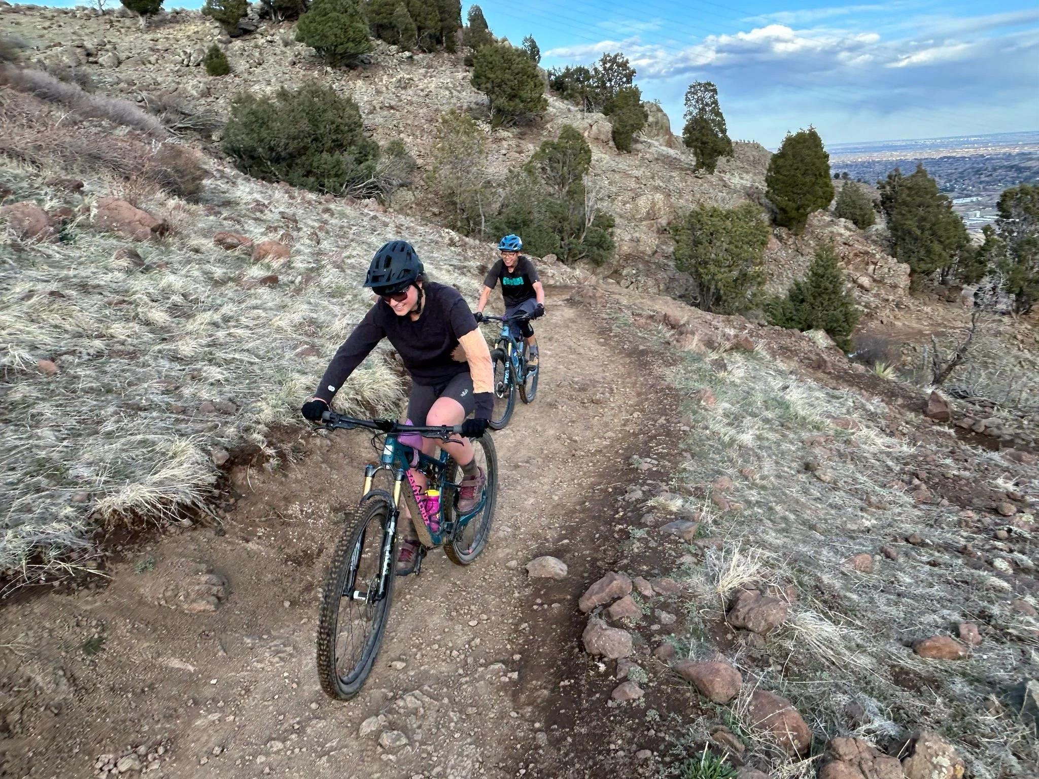 Two people mountain biking uphill on a dirt trail through a hilly, rocky landscape with sparse vegetation and trees, under a partly cloudy sky.