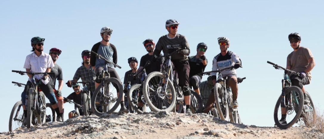 A group of people with mountain bikes on a rocky hilltop, wearing helmets and sunglasses, under a clear blue sky.