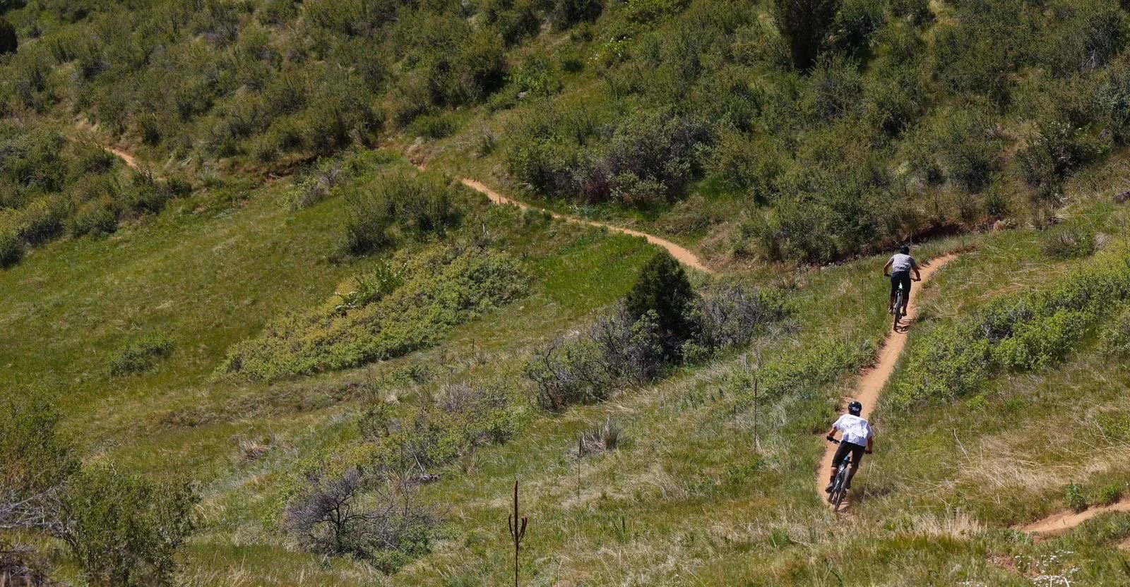 Two people riding bikes on a narrow dirt trail through green, hilly terrain with bushes and small trees.