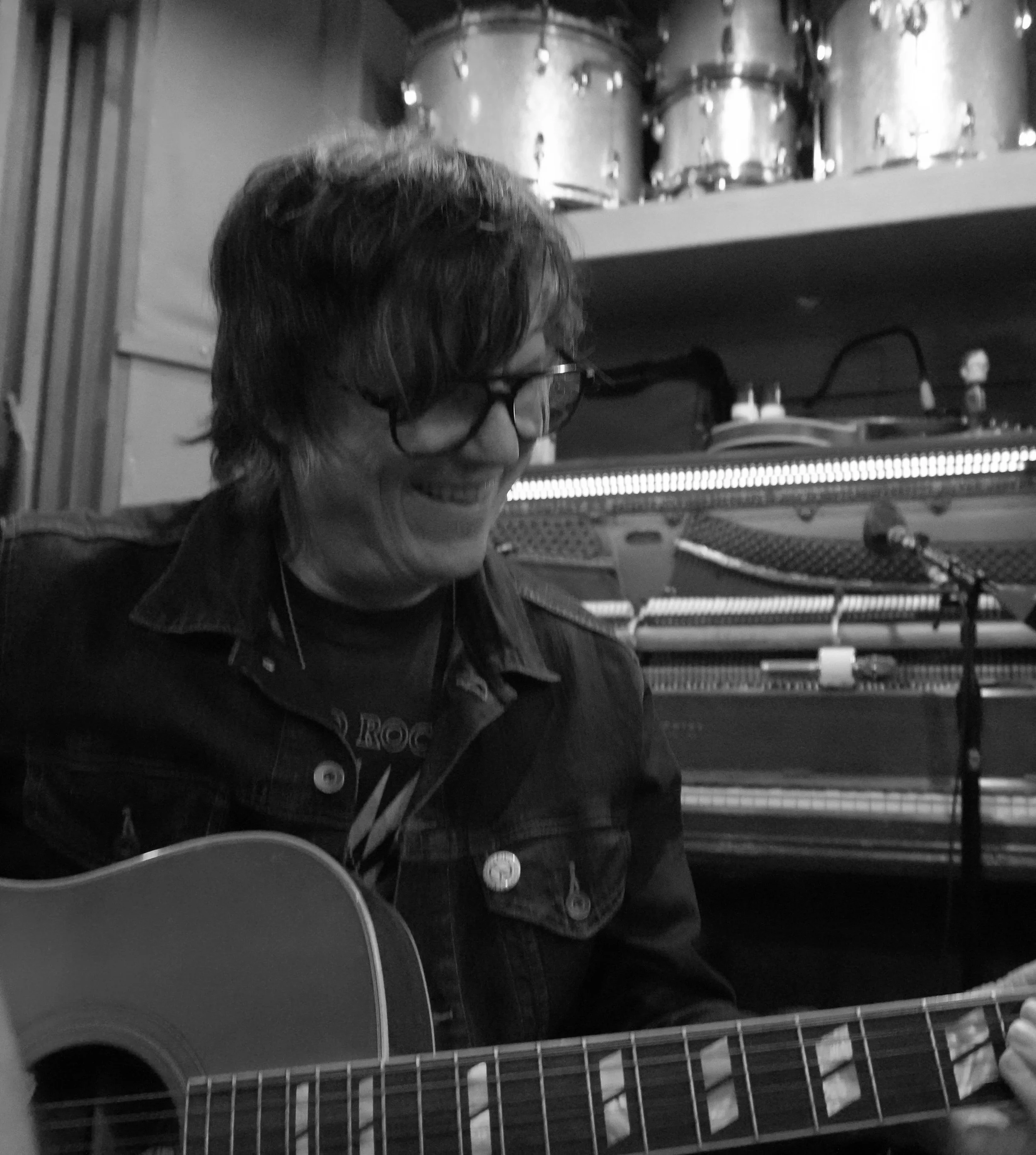 A person with glasses and shoulder-length hair plays an acoustic guitar. They are smiling and sitting in front of a piano and drum set in a music studio or rehearsal space.