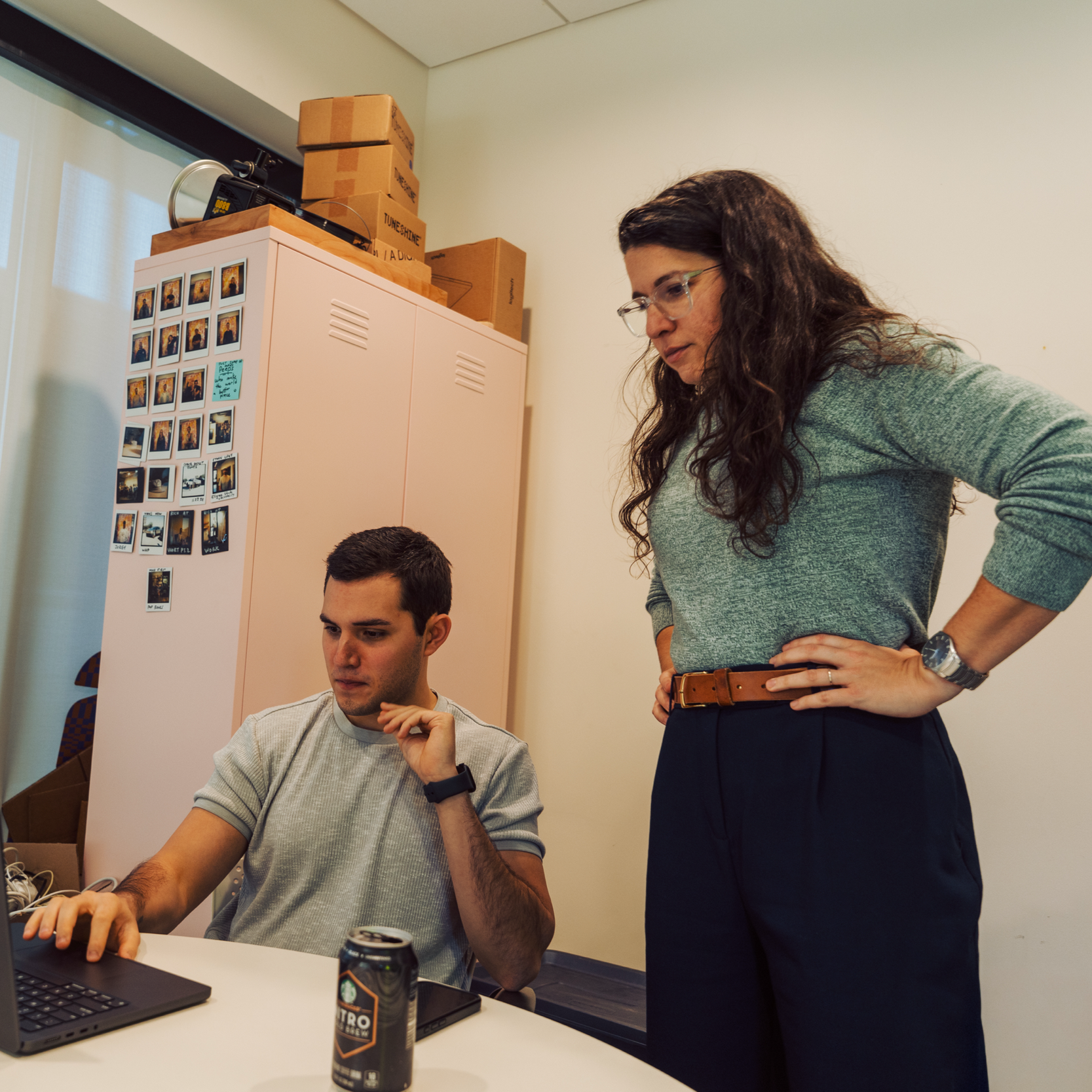 A woman and a man in an office, with the woman standing with her hands on her hips looking at the man who is sitting at a desk working on a laptop.