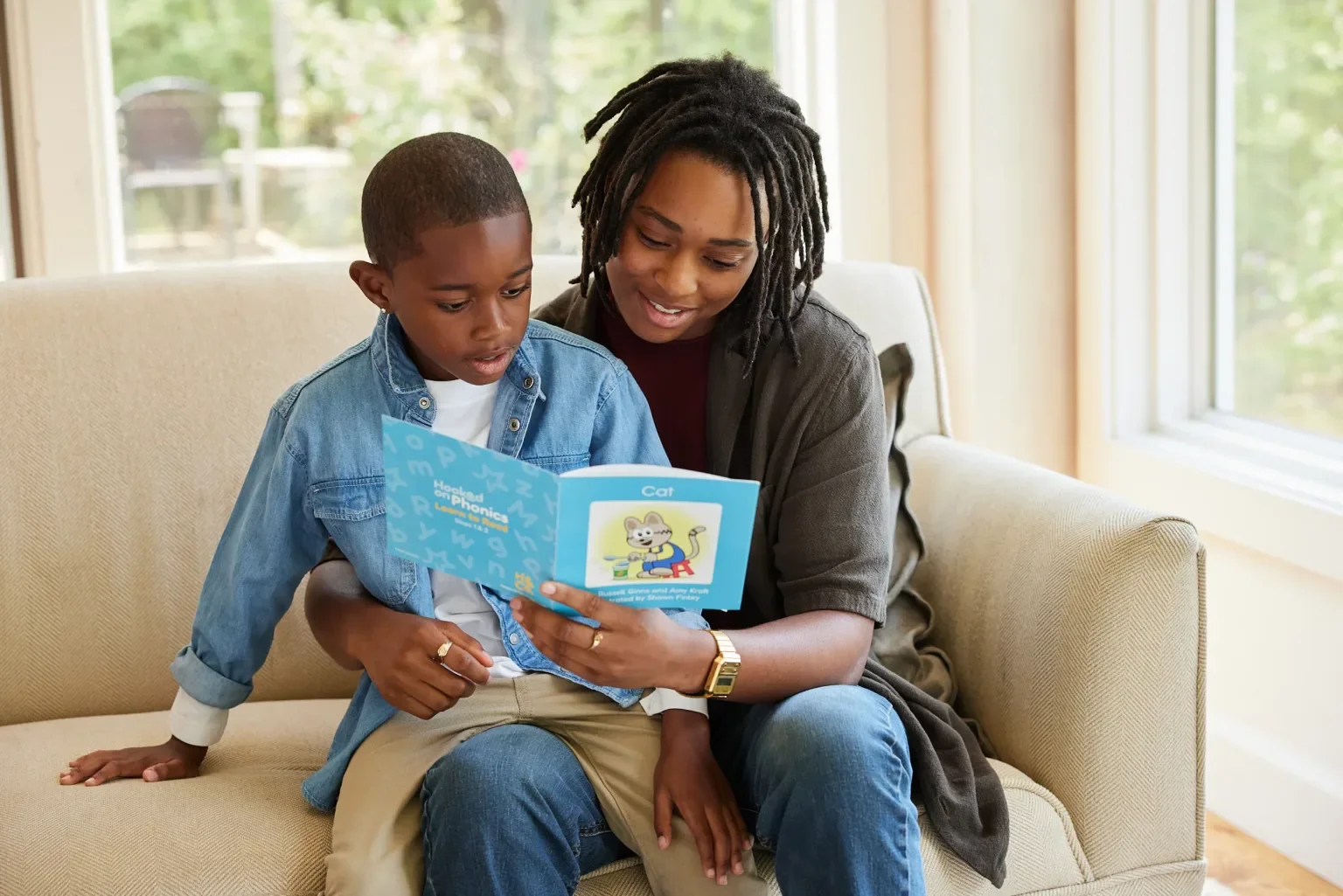 A woman and a young boy sitting on a beige sofa, reading a children's book titled 'Mouths of Phonics' with a picture of a cat on the cover, in a well-lit room with large windows and green outdoor scenery in the background.