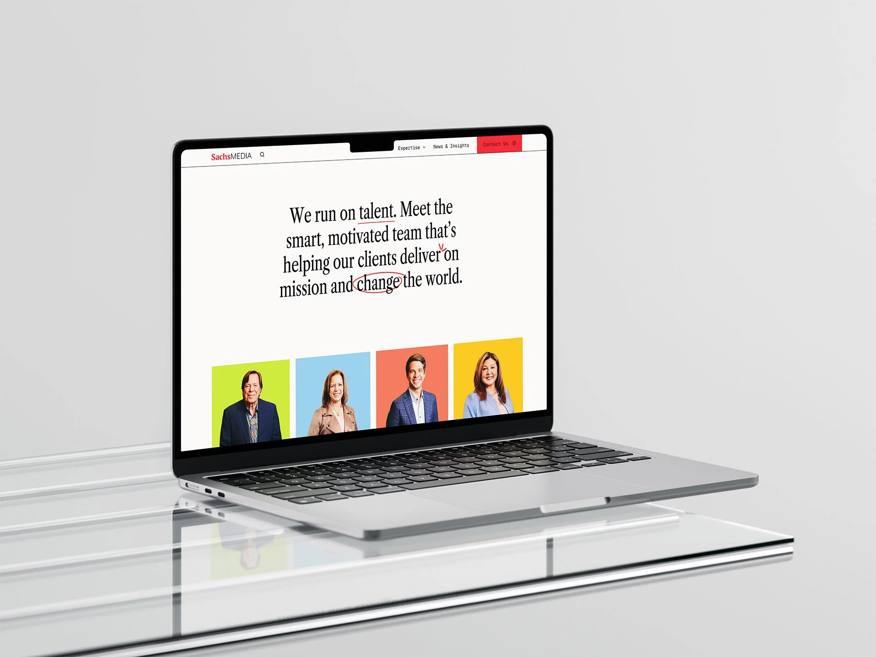 A silver laptop on a glass table displaying a website with people photos and text, against a plain white background.