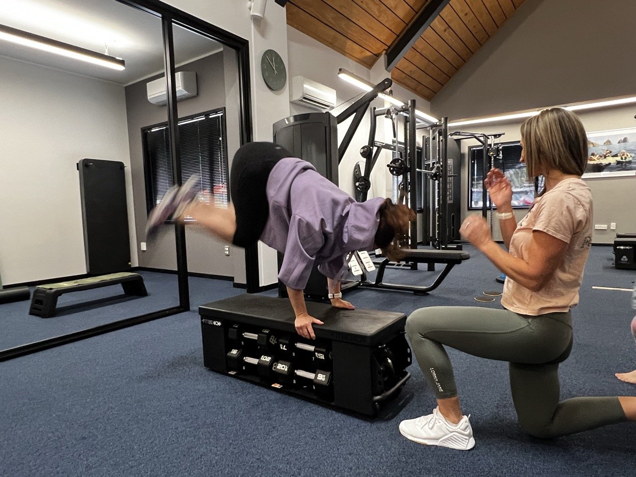 A woman in purple workout clothes is doing jump overs on a padded box while the trainer Janine Couchman observes and encourages her in a gym.