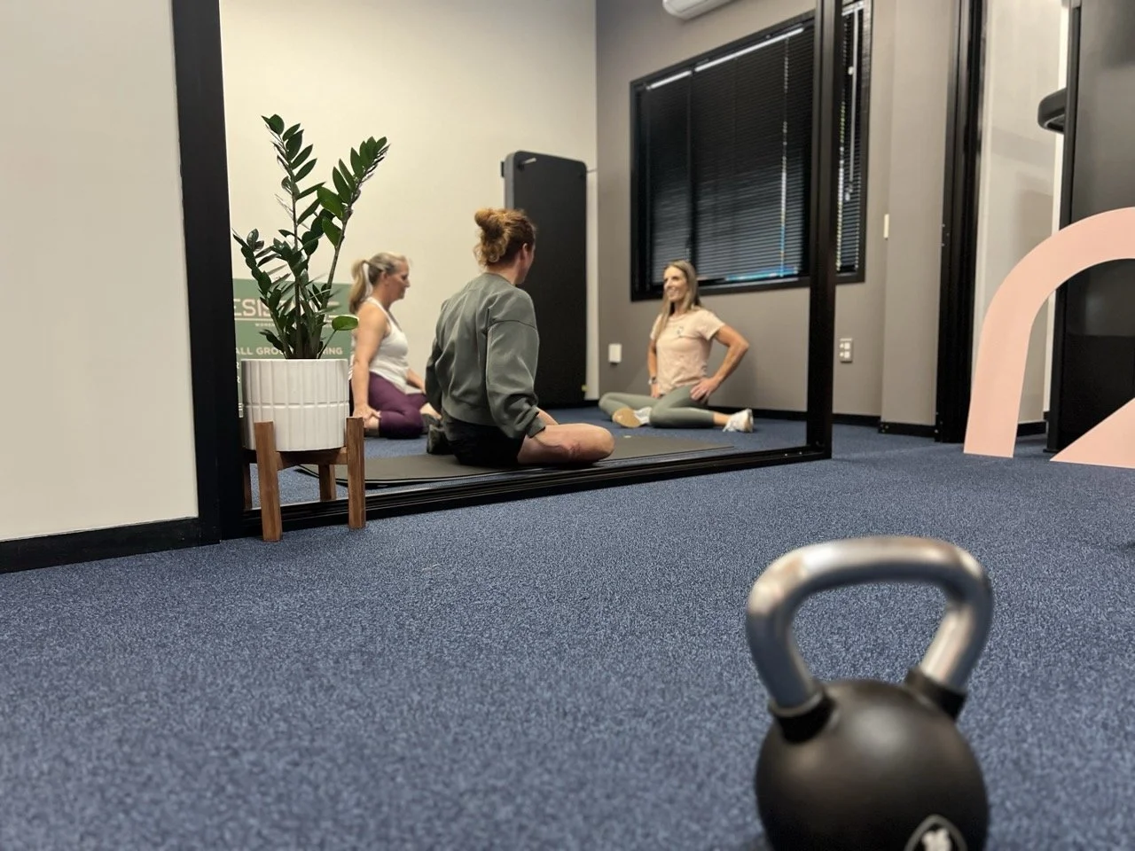 Women participating in a seated stretch with personal trainer Janine Couchman at The Resistance gym with a kettlebell on the blue carpeted floor in the foreground and a mirror reflecting the women.