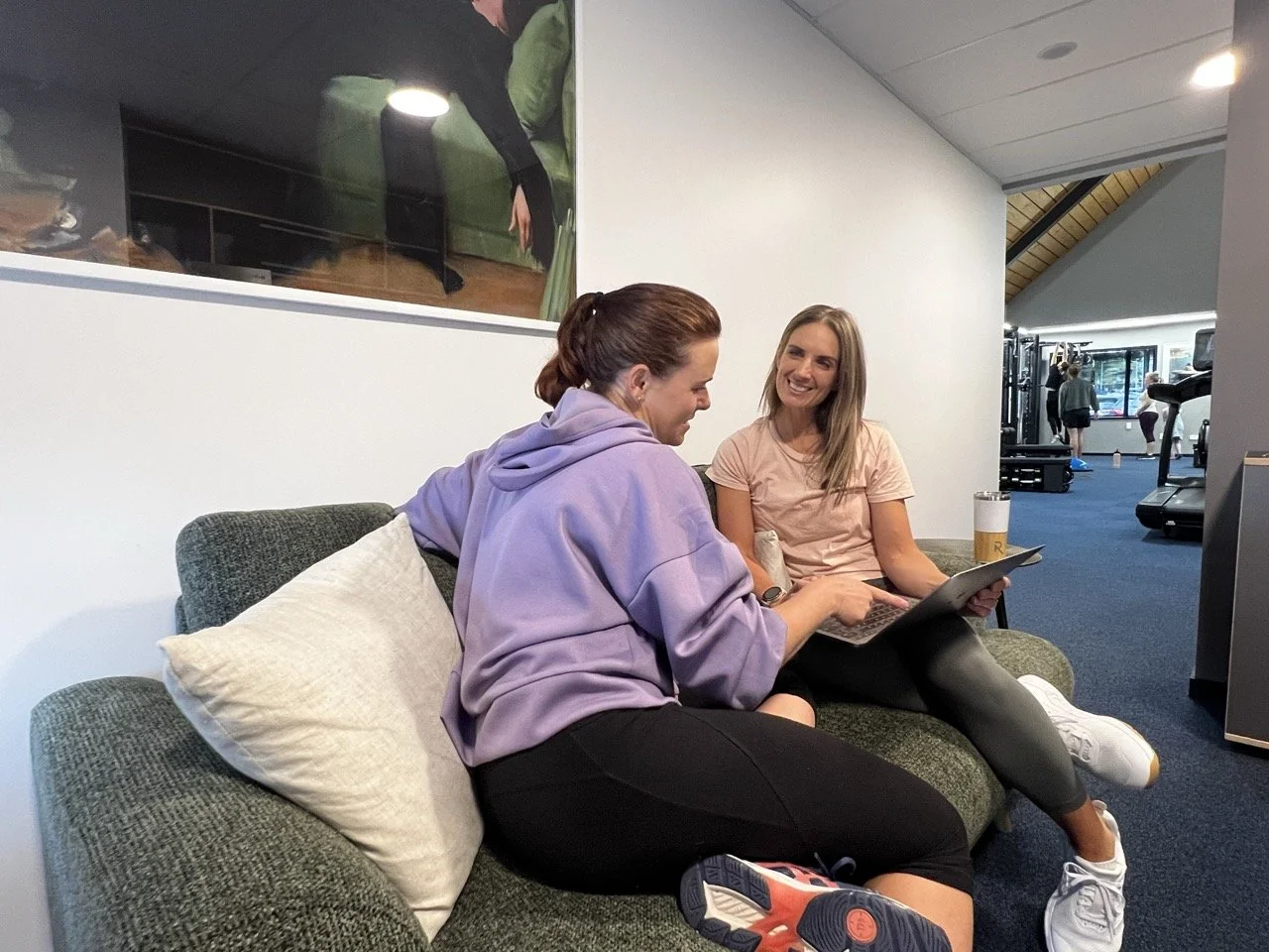 Janine Couchman sitting on a green couch with a potential gym member, smiling and looking at a laptop in a gym or fitness center lounge area. One wears a lavender hoodie, the other a pink t-shirt.