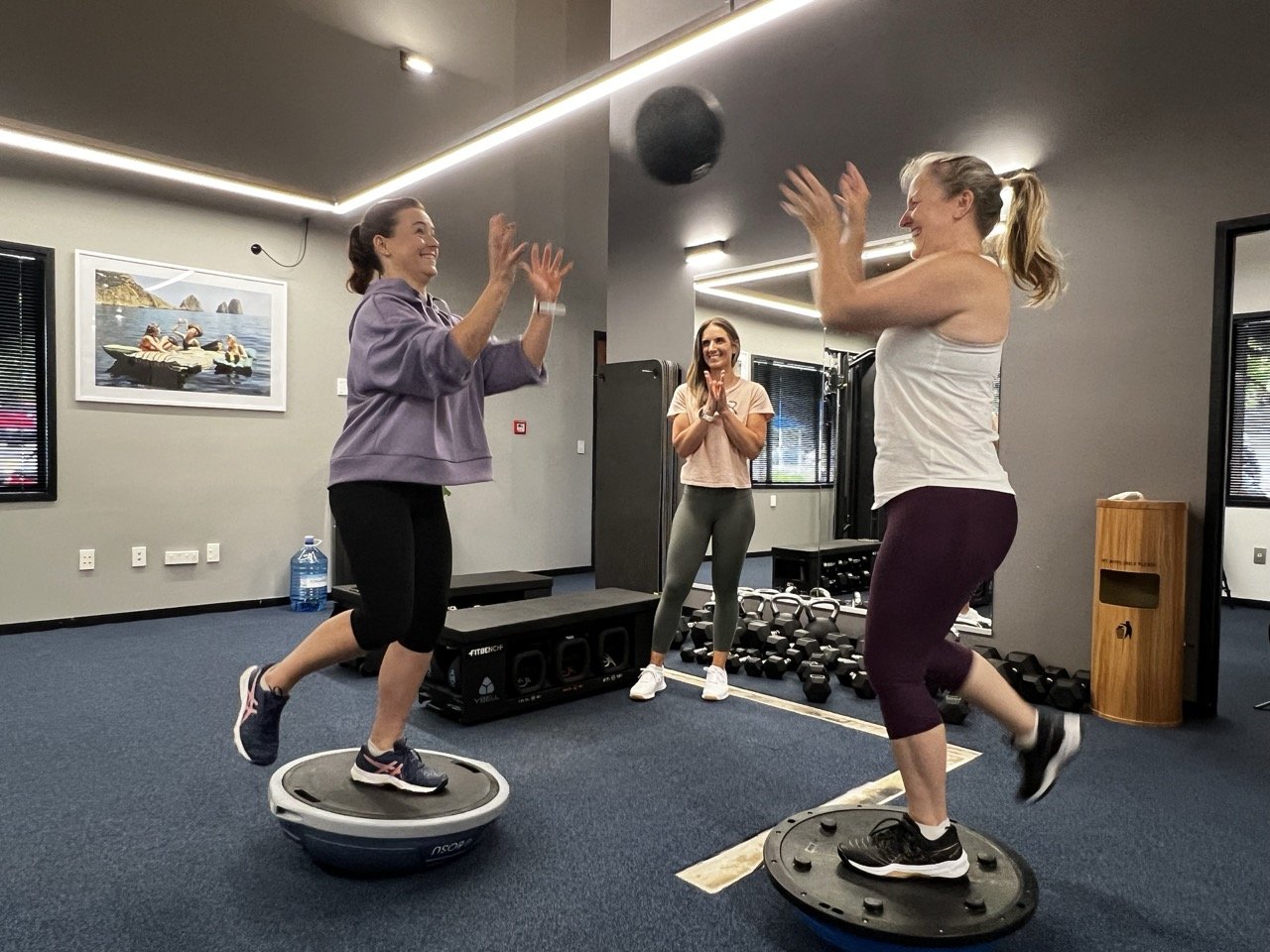 Two women on balance training discs, watched by trainer Janine Couchman, in The Resistance gym. The women on the discs are about to catch a ball. The gym has various workout equipment and a painting on the wall.