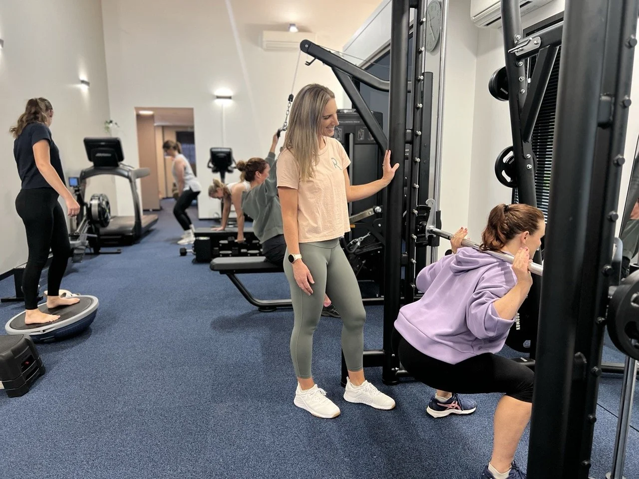 Group of women exercising in a gym, using various fitness equipment including a glider, a barbell, and a treadmill.