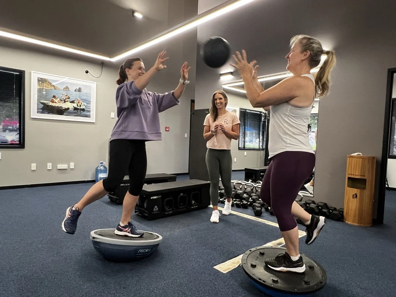 Two women are exercising on BOSU balls, passing a black medicine ball to each other, with a fitness instructor observing in the background in a gym setting.