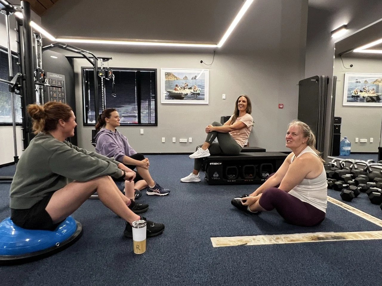 Four women sitting in a circle at The Resistance gym, engaging in a group discussion after their workout. One woman is sitting on a box with a smile, while the others sit cross-legged or on balance equipment.