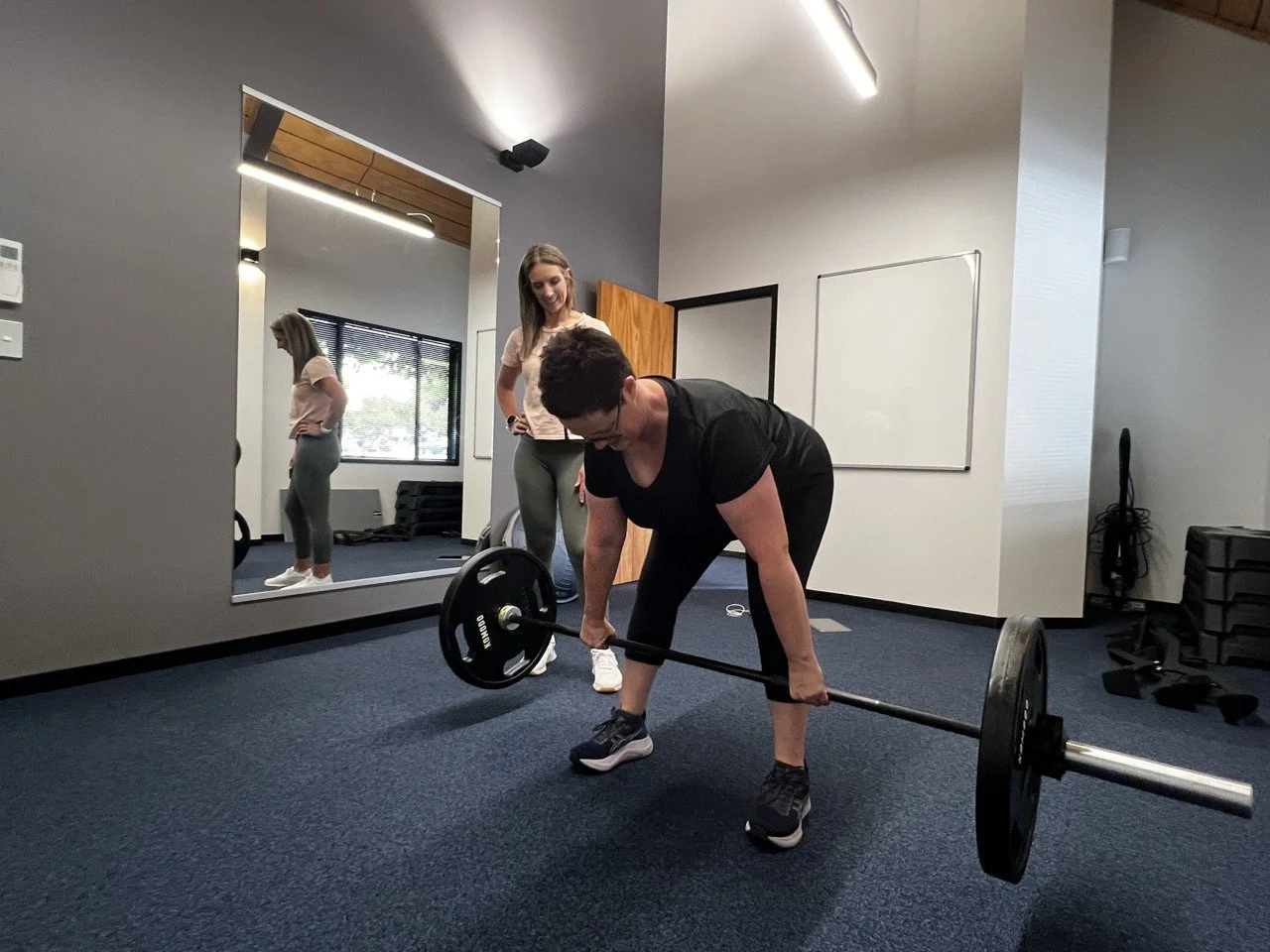 Woman lifting a barbell in a gym while personal trainer Janine Couchman observes and provides guidance, with a large mirror reflecting her.