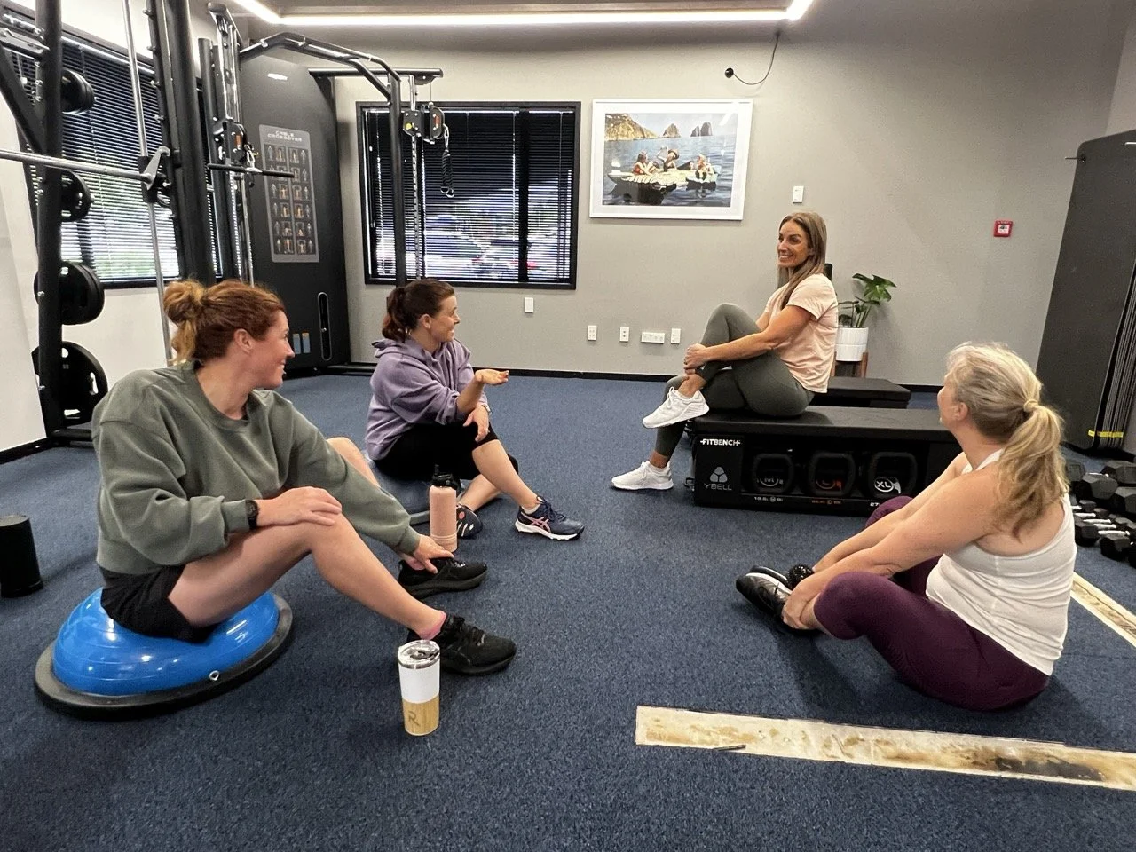 Group of women in a gym, sitting and talking during a fitness class or workout session.