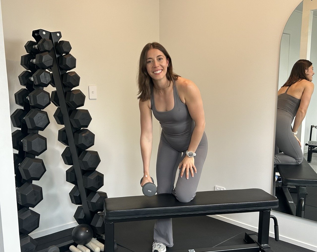 The Resistance coach and personal trainer Lilli Woodham smiling in workout attire, kneeling on a black exercise bench, holding a dumbbell in her right hand, in a home gym with black dumbbell rack and mirror reflection.