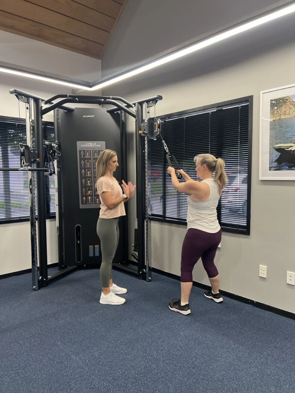Personal trainer Janine Couchman coaches a women gym member holding resistance bands attached to a cable machine, demonstrating an exercise.