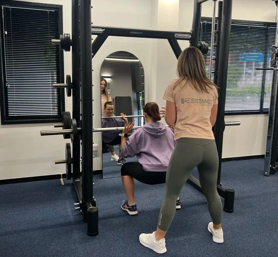 Two women at The Resistance gym with one performing a squat exercise while the other spotter, personal trainer Janine Couchman, stands behind her, all looking into a mirror.