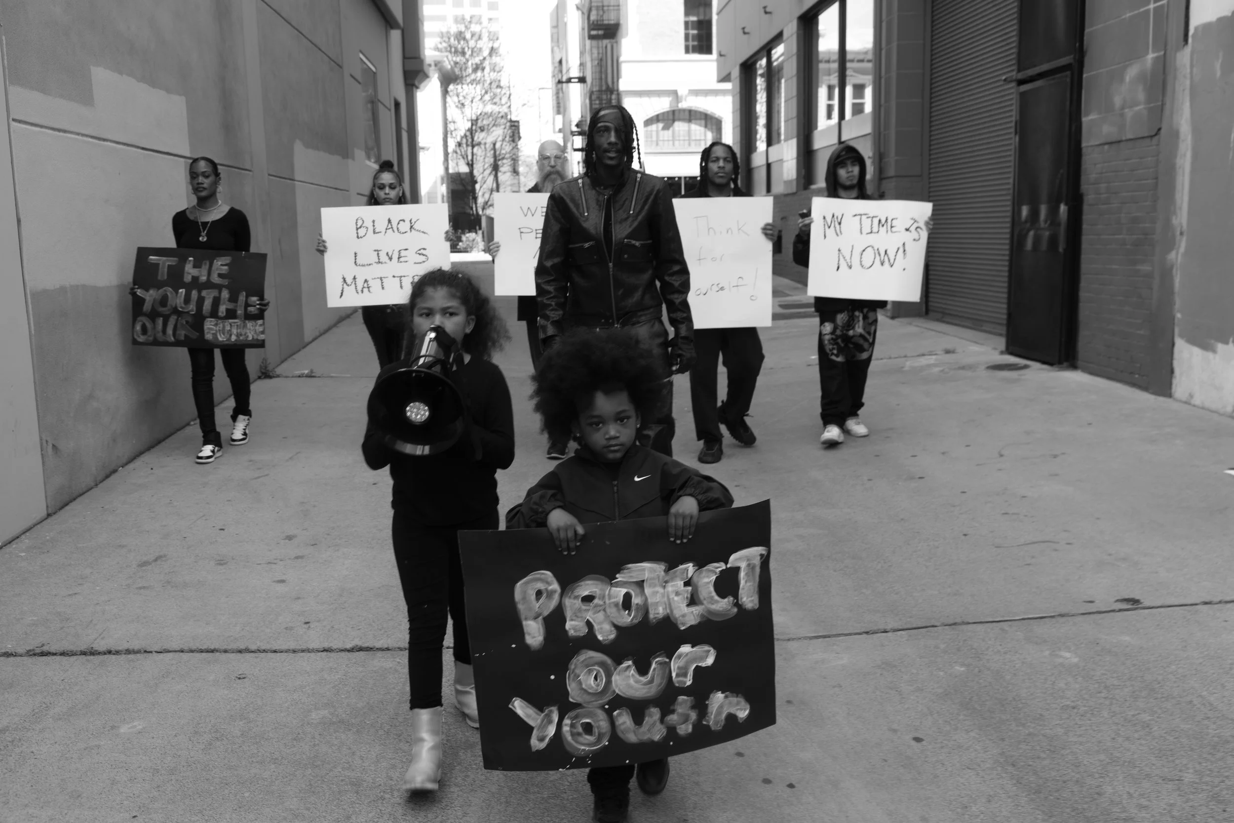 A diverse group of people, including children, protesting on a city street holding signs with messages such as 'Protect Our Youth,' 'Black Lives Matter,' 'Think for Yourself,' 'My Time Is Now,' and 'The Youth Our Future.'
