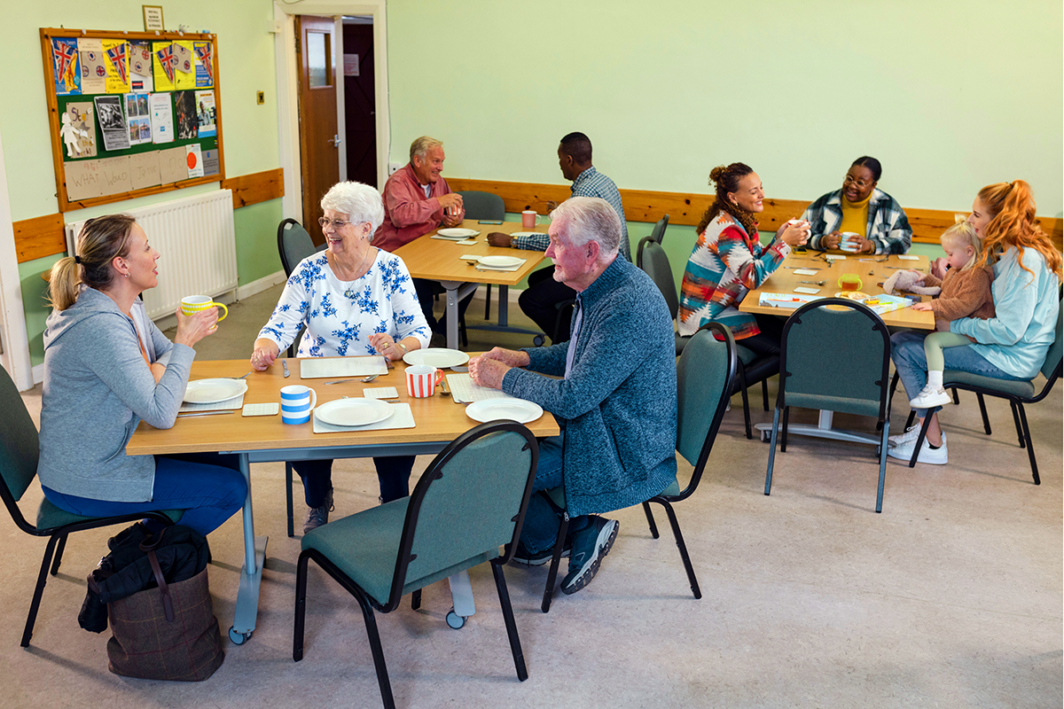 A group of elderly and young people sitting at tables, chatting and enjoying a meal together in a community room with lime green walls and colorful decorations.