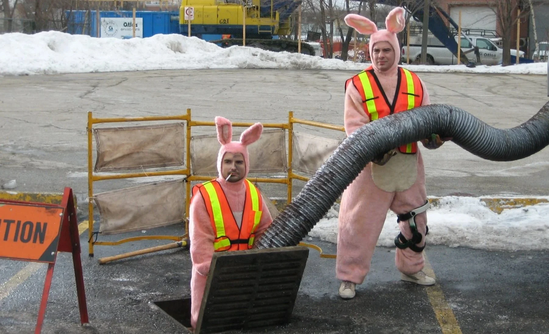 Two men dressed in pink bunny costumes, one standing and one sitting, at a construction site during winter. The standing man is holding a large black hose, and both are wearing reflective safety vests. There is snow on the ground and construction barriers in the background.