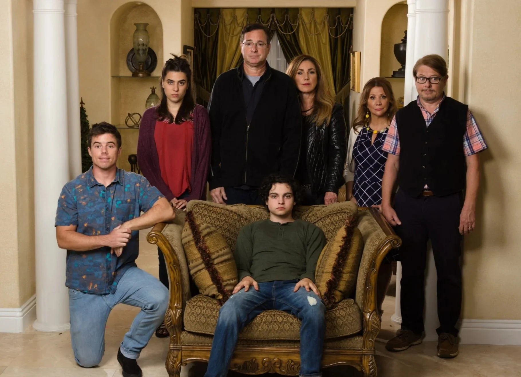 A group of eight people pose inside an elegant living room, with one person seated in an armchair and others standing behind, facing the camera with serious expressions.