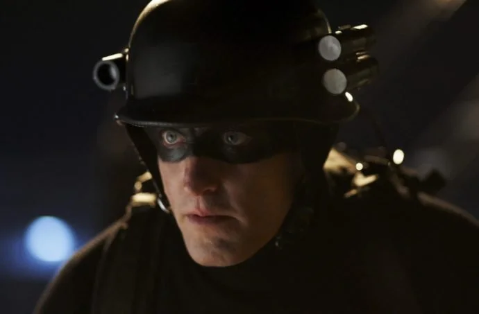 A close-up of a man wearing a black helmet and mask with mounted lights, his face lit dramatically in a dark, industrial setting, creating a tense, futuristic or sci-fi atmosphere.