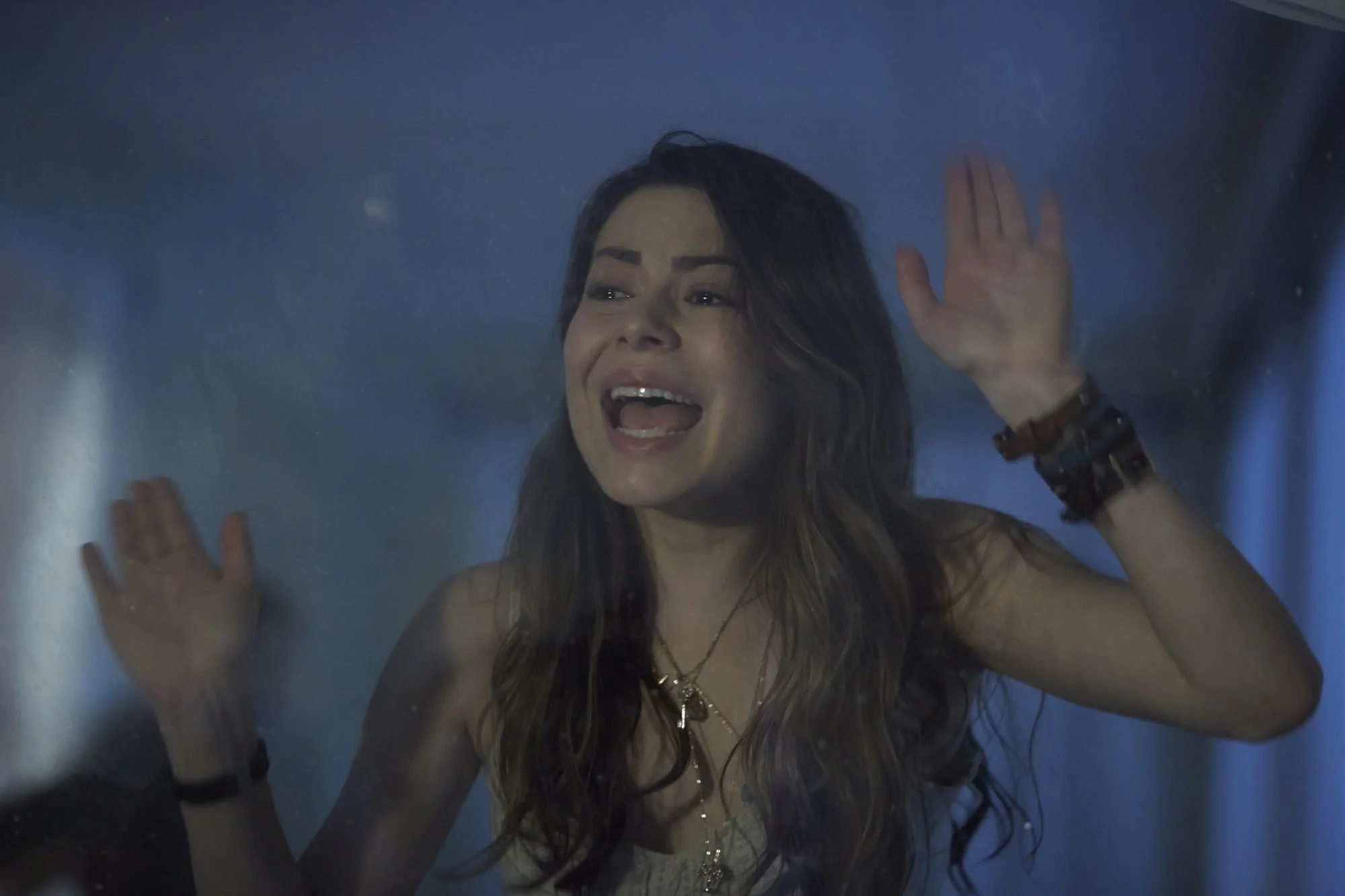 A young woman presses her hands against glass, shouting with a distressed expression, seen through a blue-toned, foggy surface.