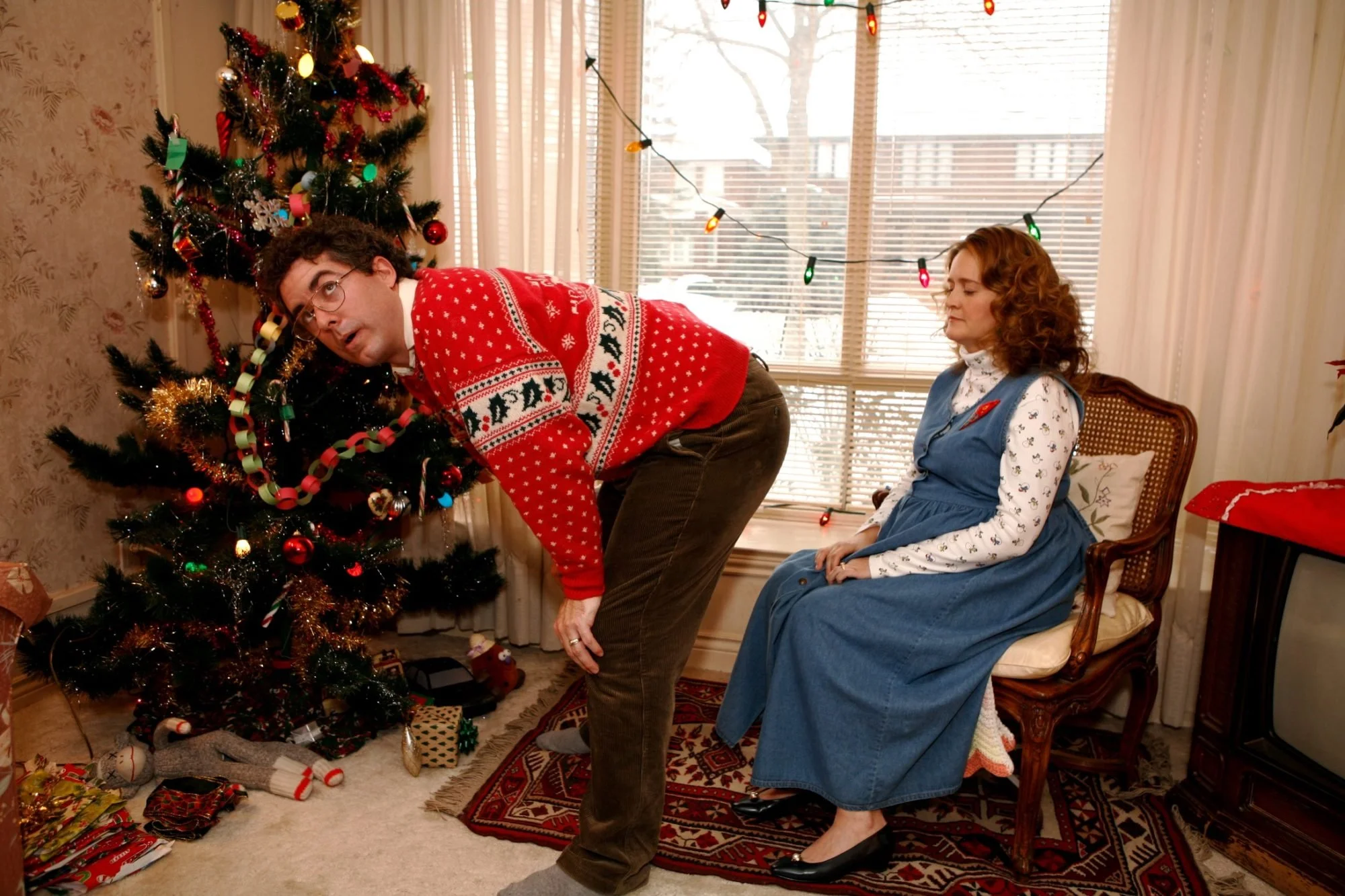A man in a festive Christmas sweater bent over near a decorated Christmas tree, with a woman sitting in a chair watching him. The room has holiday decorations and presents under the tree.