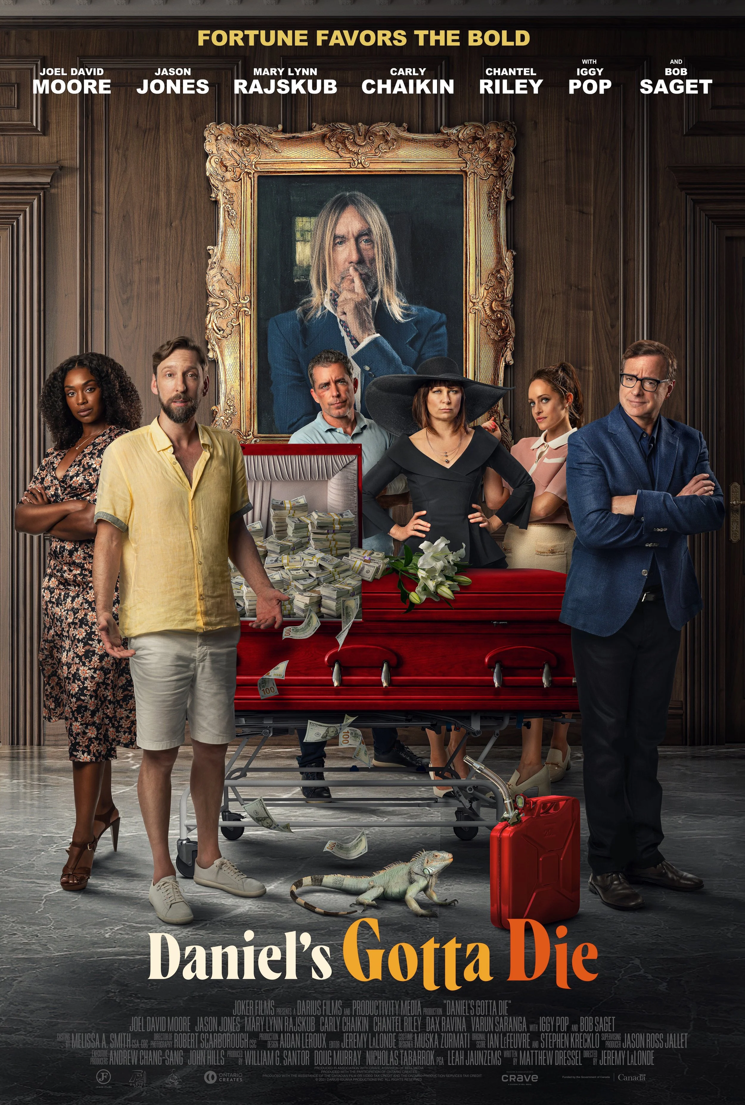 Group of seven people dressed in formal and casual attire standing around a red coffin with money scattered around it, inside a wood-paneled room with a large framed portrait of a man with long blonde hair in the background. A small lizard and a red gasoline can are on the floor.