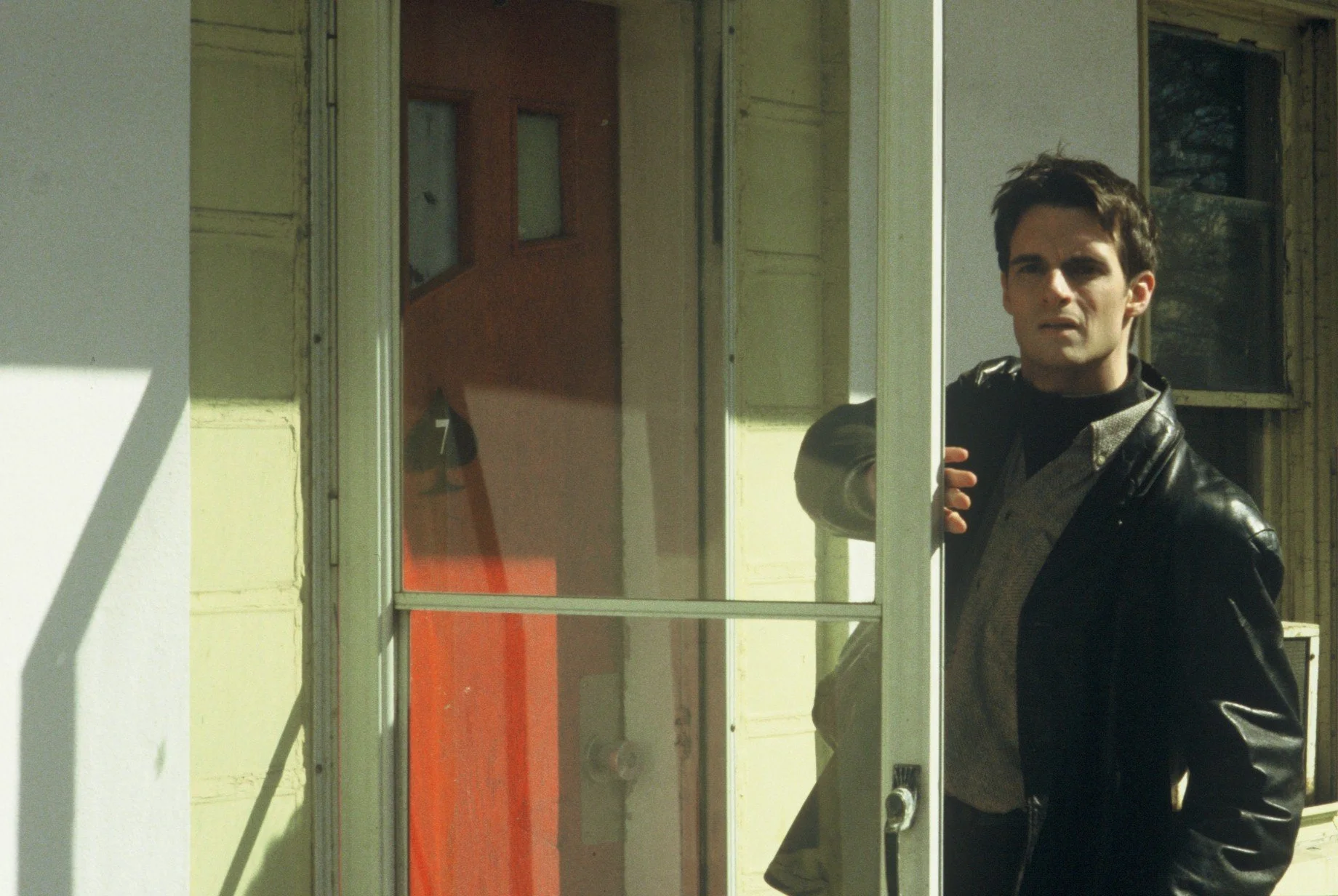 A man in a dark jacket stands in a doorway of a modest roadside motel, partially framed by a glass door, looking outward in daylight with a tense, introspective expression.