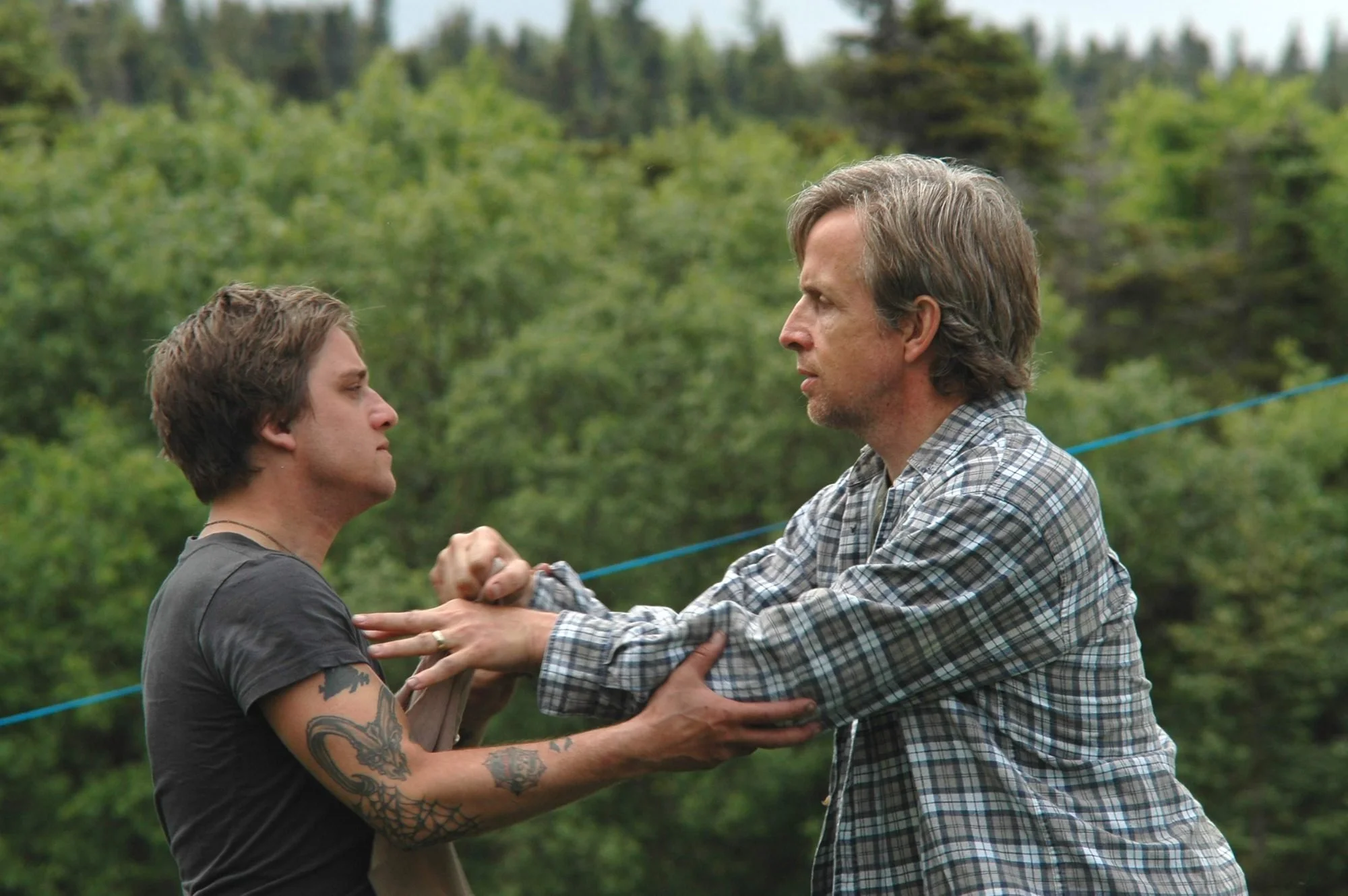 Two men engaged in a physical confrontation outdoors, with one man grabbing the other's collar and the other pushing him back. They are surrounded by green trees.