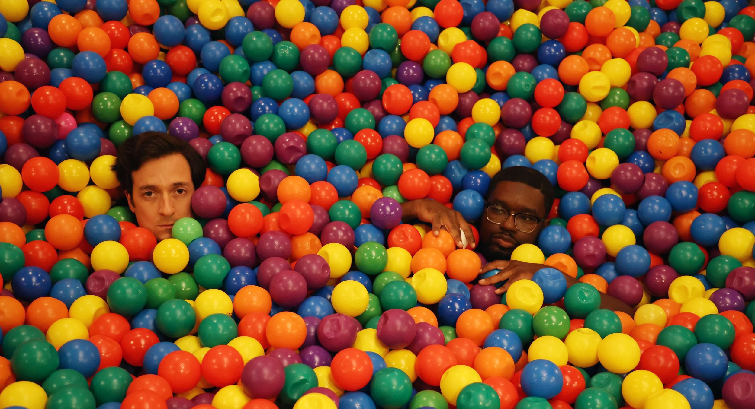 Two men partially submerged in a colorful ball pit, surrounded by bright plastic balls, looking toward the camera in a playful, surreal setting.
