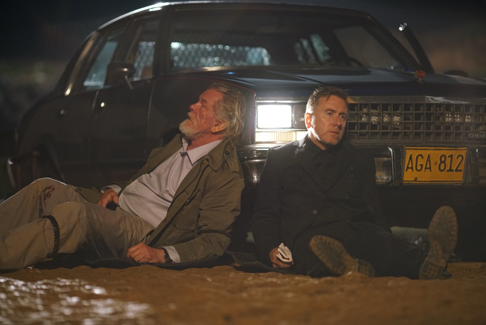 Two men sit on the ground at night, leaning against the front of a parked car with its headlights on, appearing exhausted and tense on a muddy roadside.