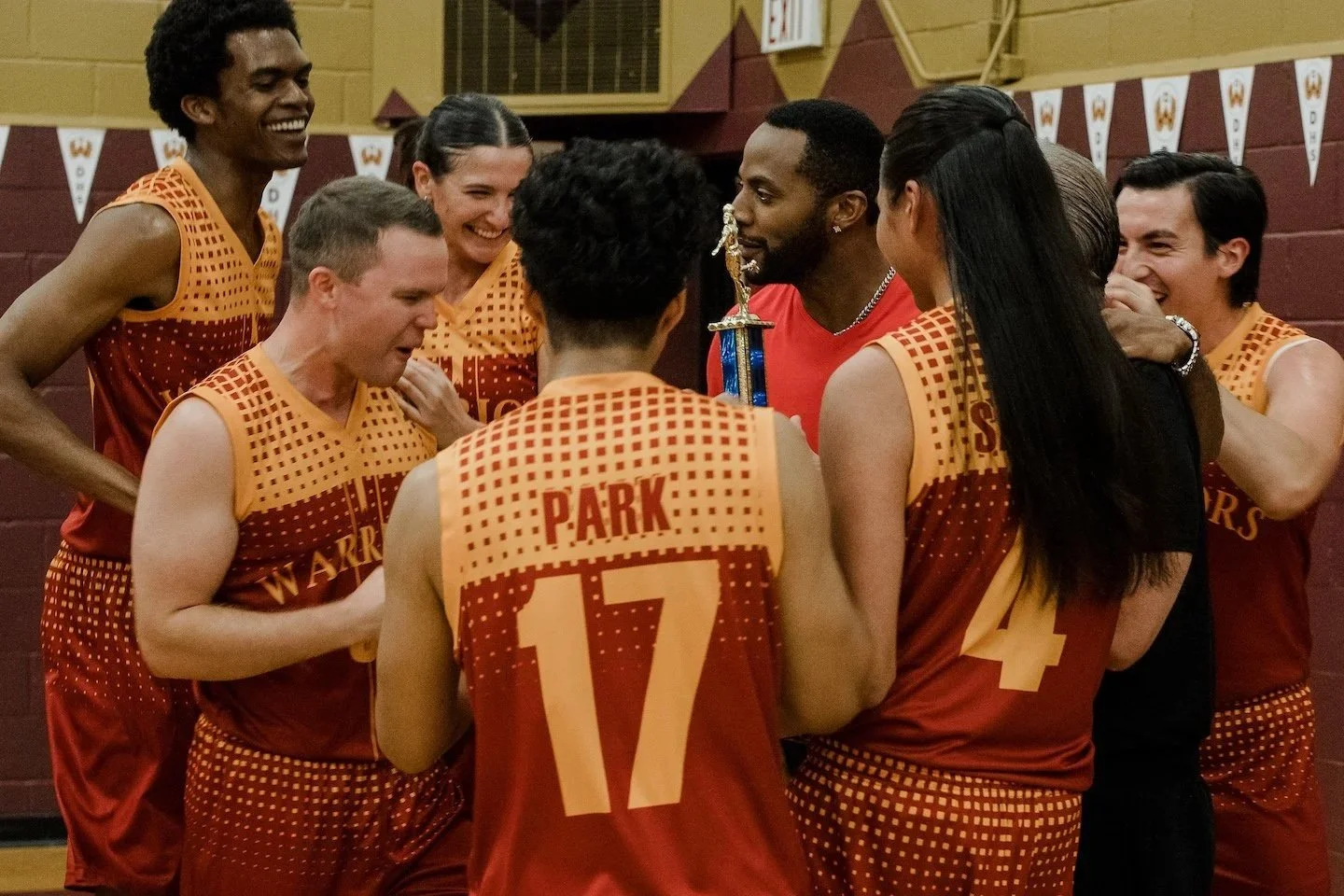 A group of basketball players in matching uniforms celebrate together in a gym, smiling and crowding around a small trophy.