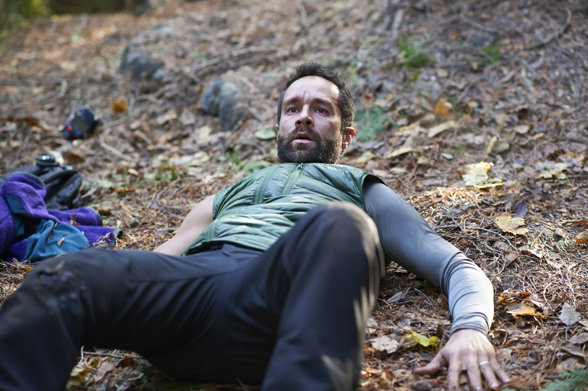 A bearded man lies on a forest floor covered in leaves, looking shaken and catching his breath, dressed in outdoor clothing in a wooded setting.