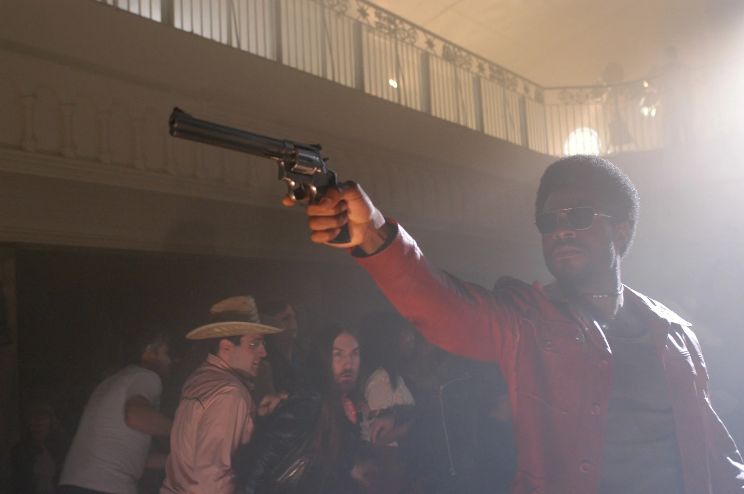 Man in red jacket and sunglasses aiming a revolver inside a smoky indoor crowd, dramatic cinematic scene