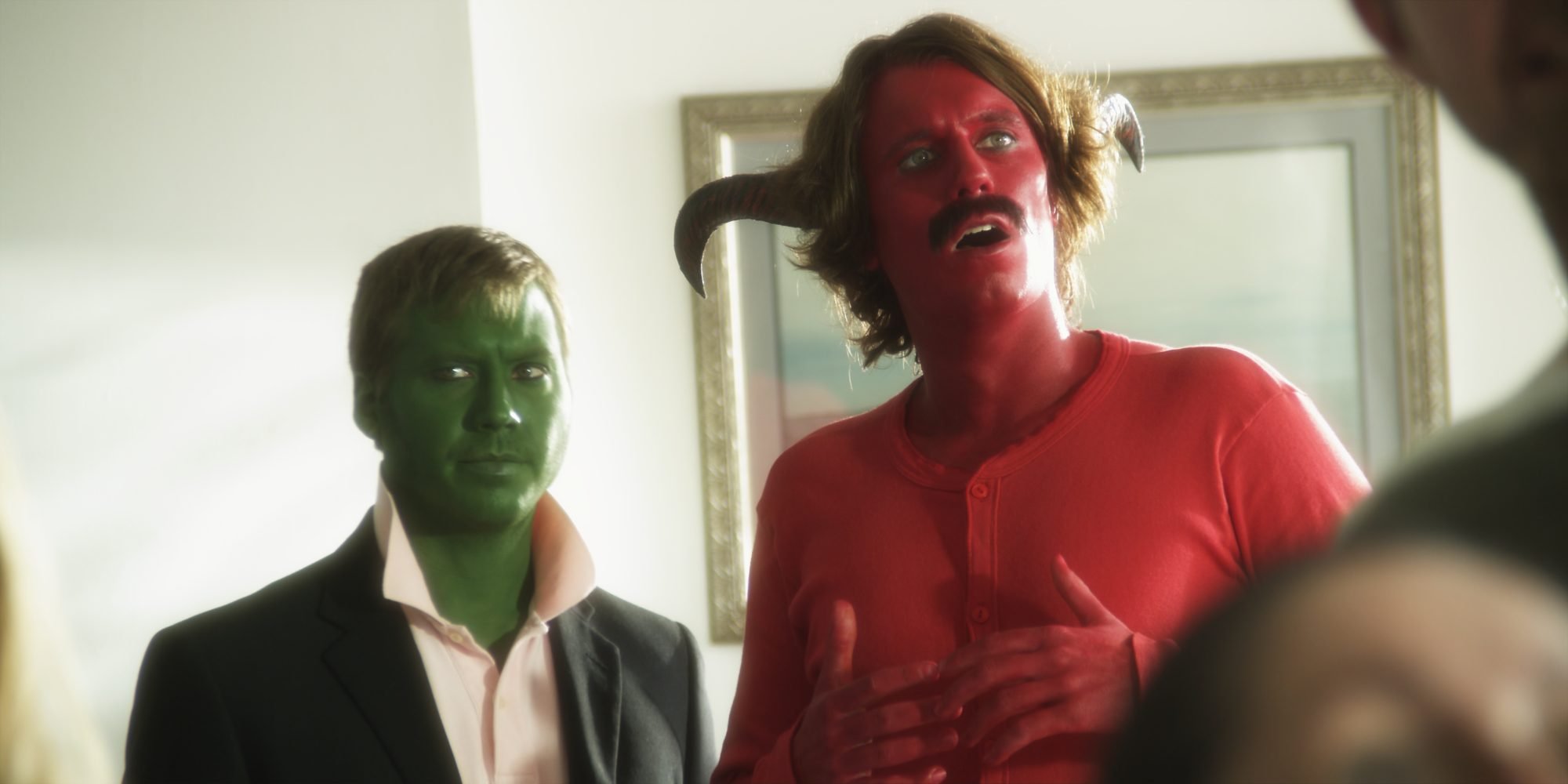 Two men in theatrical makeup stand indoors: one with green-painted skin wearing a suit, and the other with red-painted skin, small horns, and a mustache, gesturing mid-speech, creating a surreal, comedic, and fantastical scene.