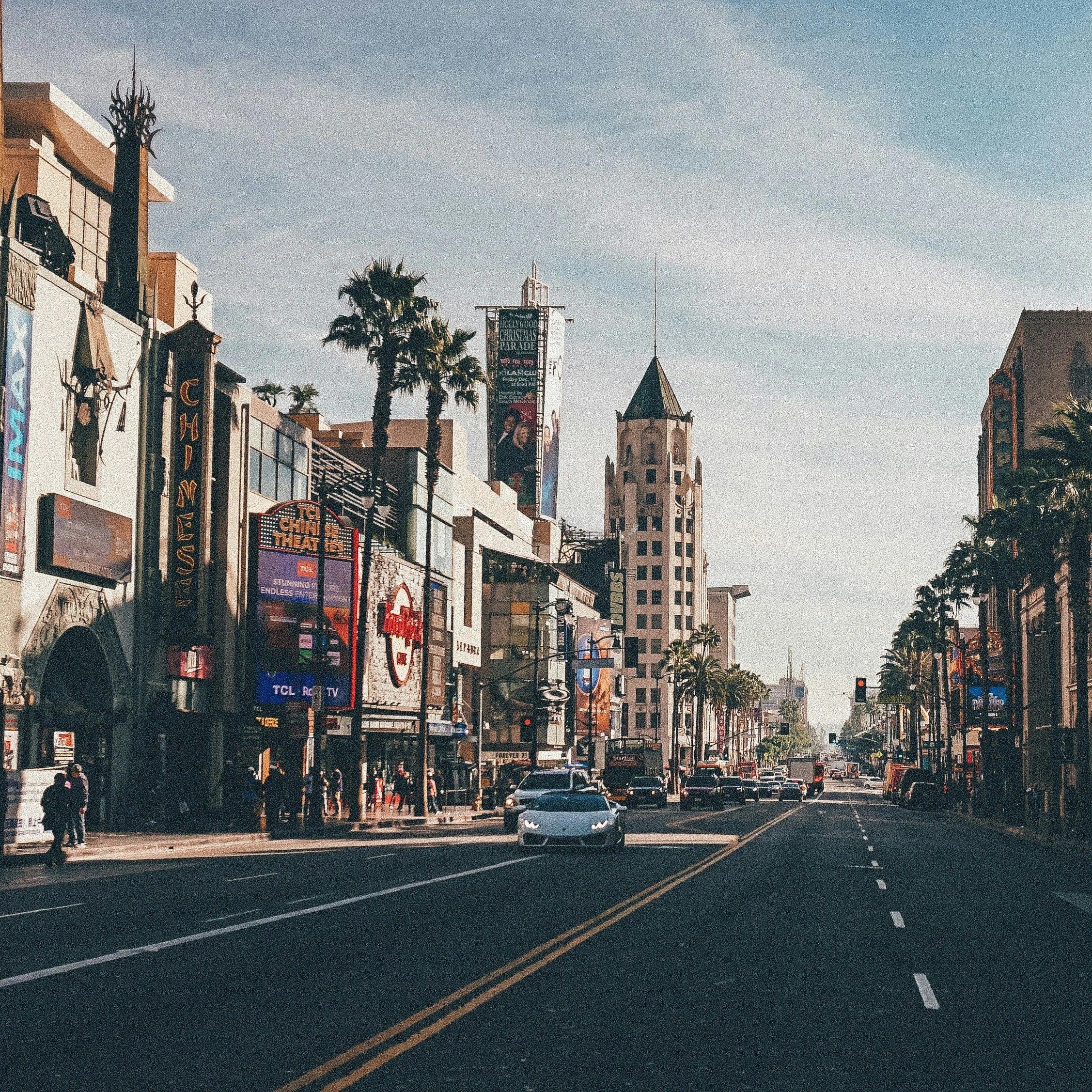 Street scene featuring shopping and entertainment venues, palm trees lining the sidewalks, cars on the road, and a tall, pointed building in the background, likely in Los Angeles.