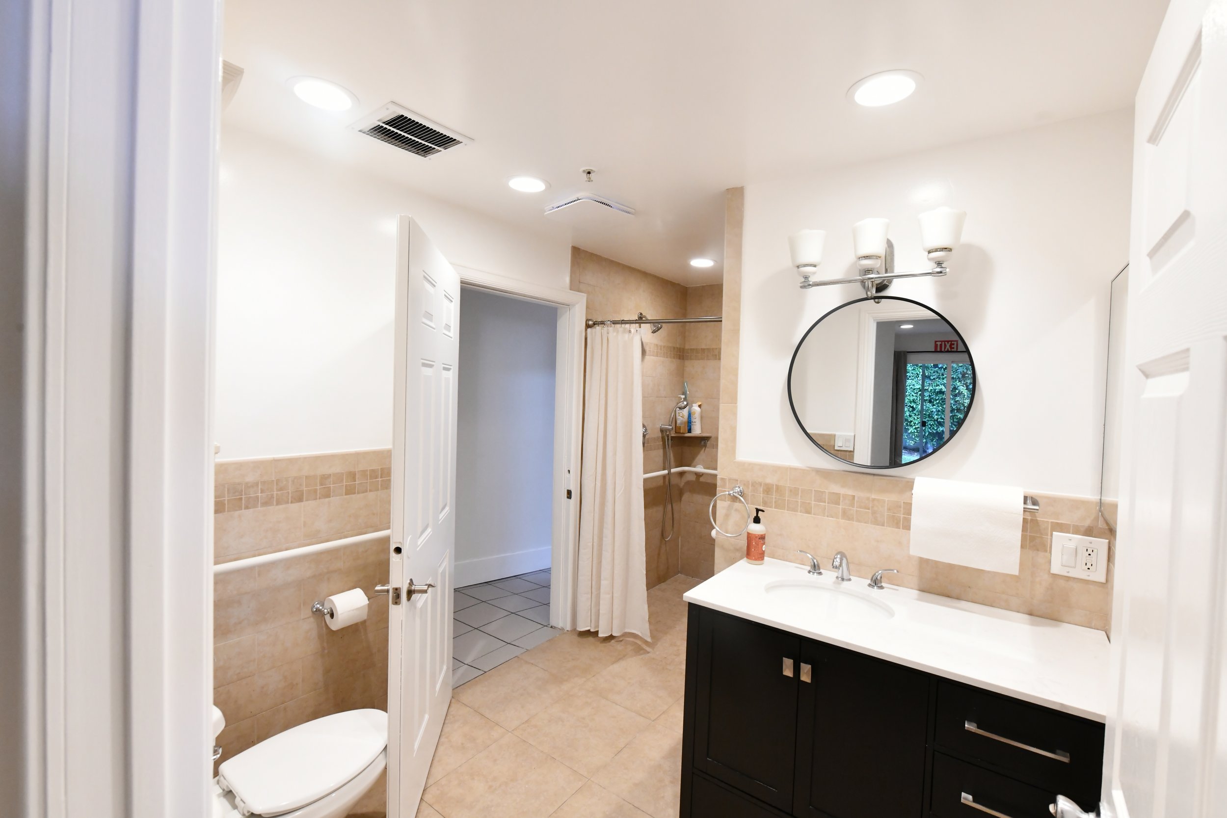 Bathroom with a separate toilet area, a shower with curtain, a vanity with a round mirror, double sinks, and wall-mounted light fixtures.