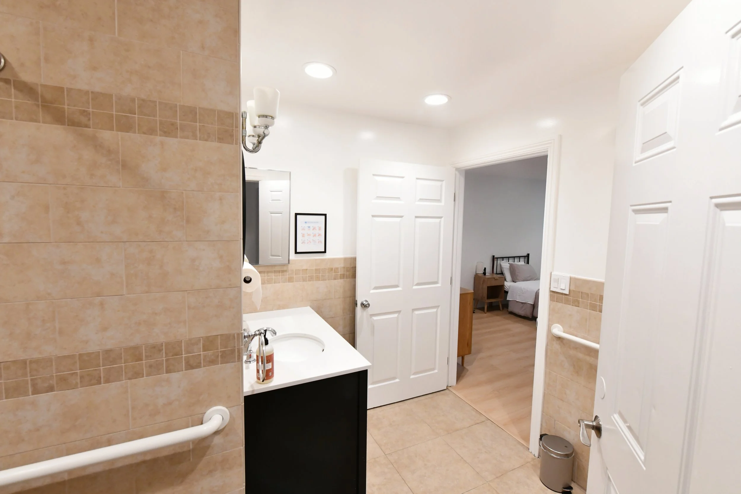 A bathroom with beige tiles, a white ceiling with recessed lighting, a mirror, a black vanity with a white countertop, and a door leading to a bedroom with a bed and nightstand.