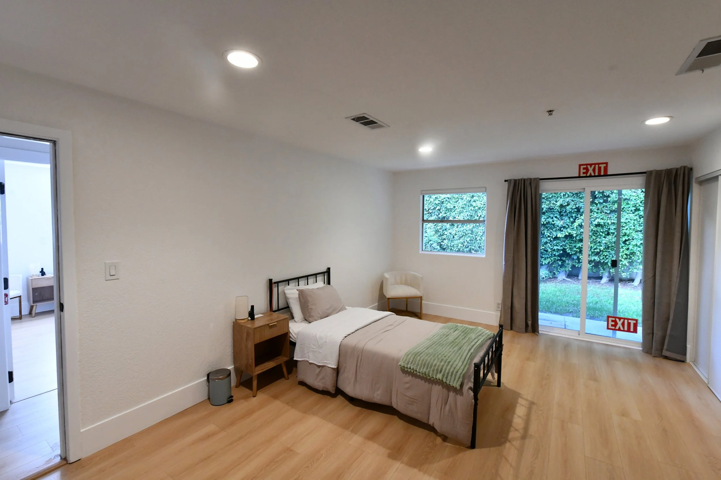 A bedroom with a single bed, nightstand, beige walls, hardwood floor, a white chair near window, sliding glass door with curtains and "EXIT" signs, outside green foliage visible.