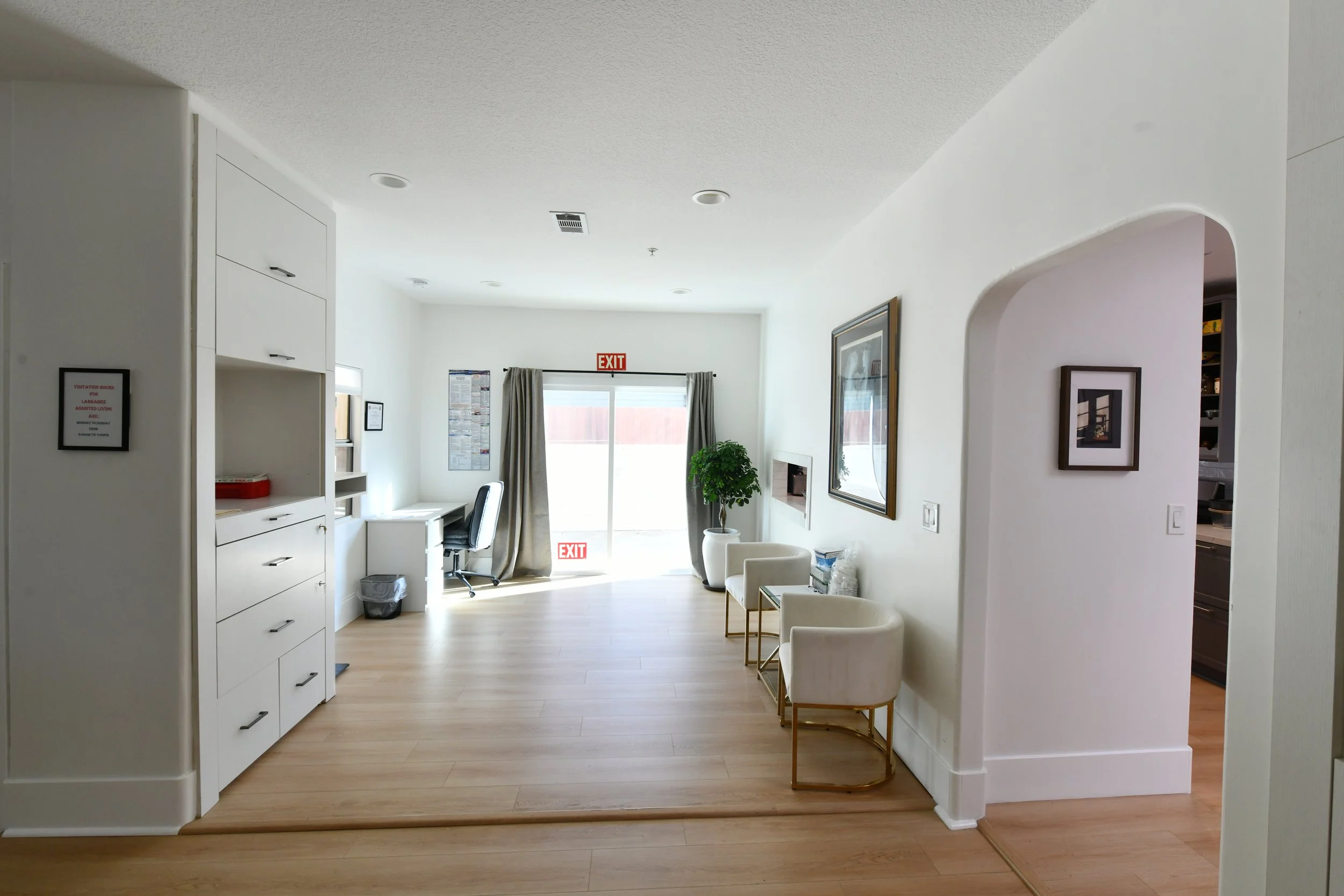 Interior of a bright, modern room with white walls, light wood flooring, a desk with a chair near the sliding glass door, and seating area with two white armchairs and a small table.