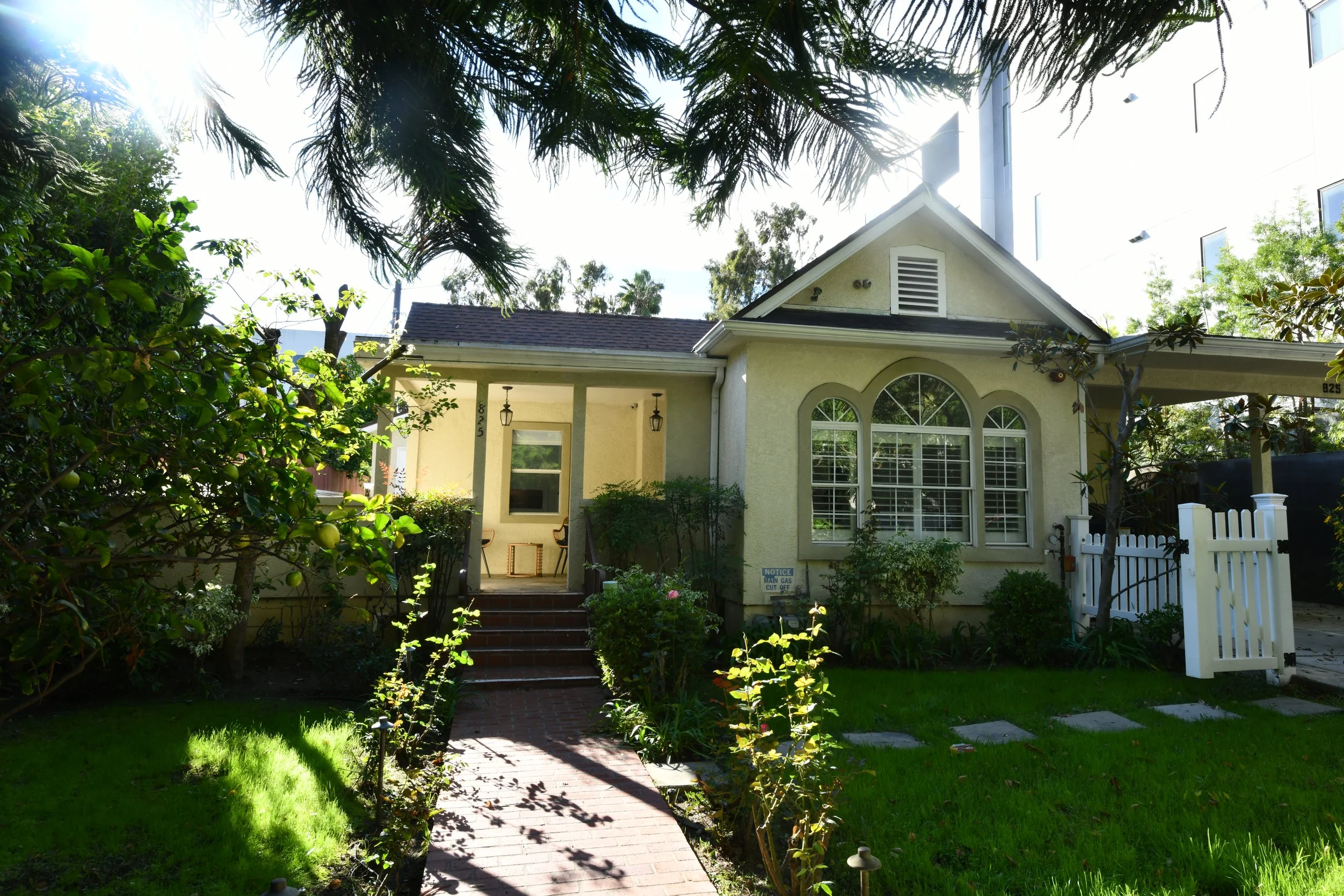 A single-story house with a front yard, green lawn, and a brick pathway leading to the front porch. The house has large arched windows, a small porch with two chairs, and is surrounded by trees and bushes. Bright sunlight and part of a tree canopy ar