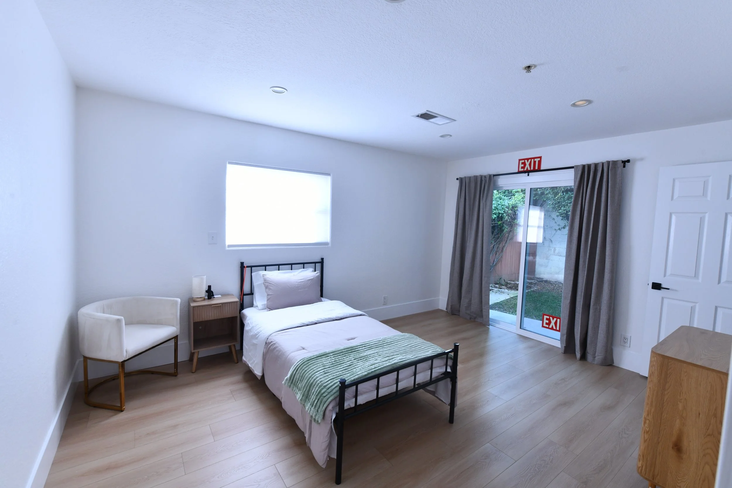 A minimalist bedroom with a bed, white walls, wood flooring, a nightstand with a lamp and phone, a white chair, a window, and a sliding glass door with curtains leading to an outdoor area.
