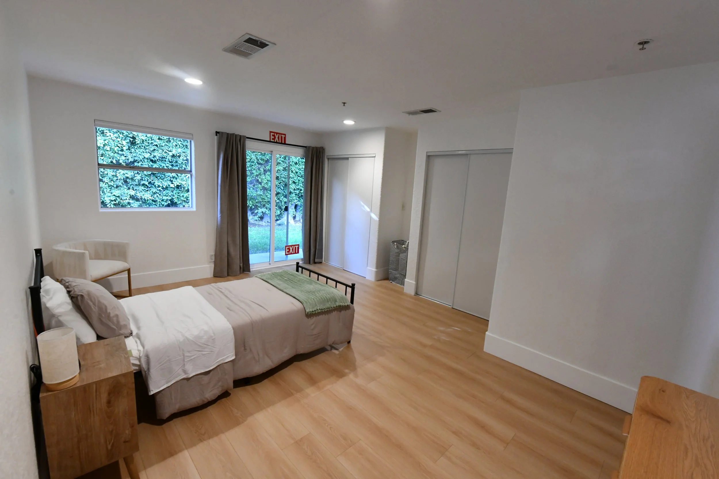 A bedroom with a single bed, a wooden nightstand with a lamp, a beige armchair near the window, a sliding glass door with gray curtains, and a closet with sliding doors. The room has hardwood flooring and white walls.