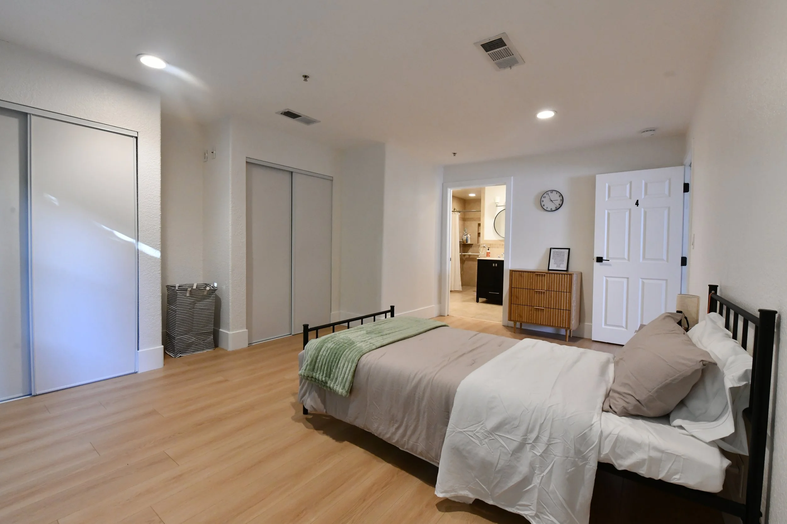 A bedroom with a bed, closet, and an adjoining bathroom, with wooden flooring and white walls.