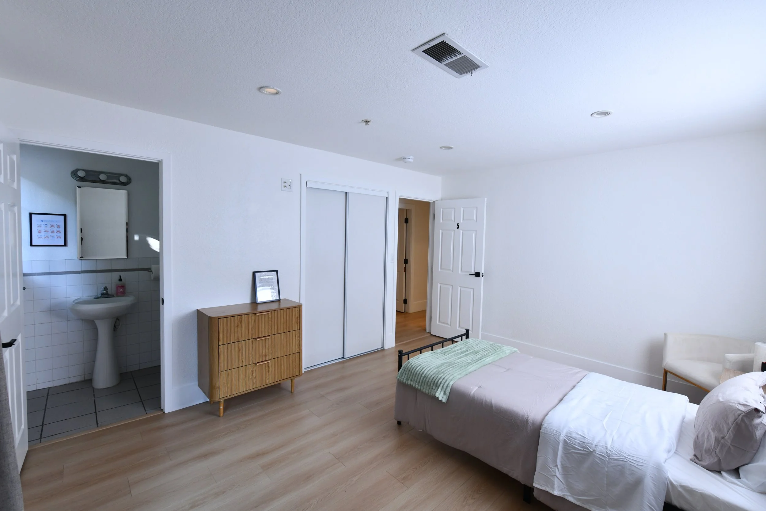 A minimal bedroom with a twin bed, a wooden dresser, a chair, and an open door leading to a bathroom. The bathroom has a pedestal sink and a mirrored medicine cabinet. The room has white walls, wooden flooring, and ceiling air vents.