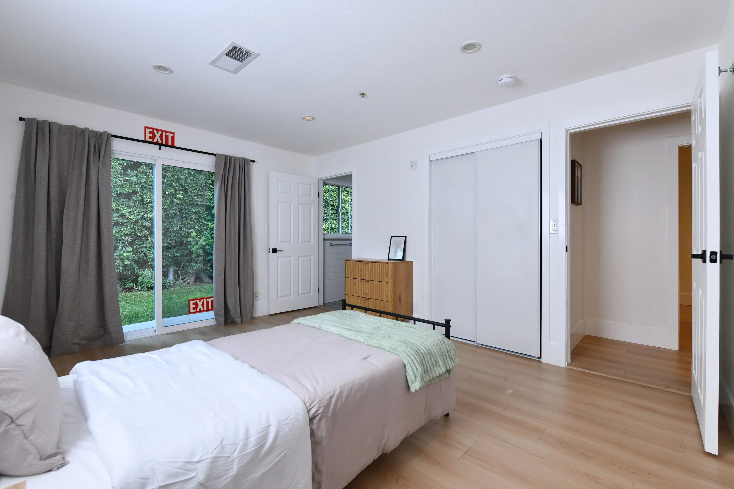 Bright bedroom with a bed, sliding glass door with outdoor greenery, gray curtains, wooden dresser, closet with sliding doors, and a partially open door leading to a hallway.