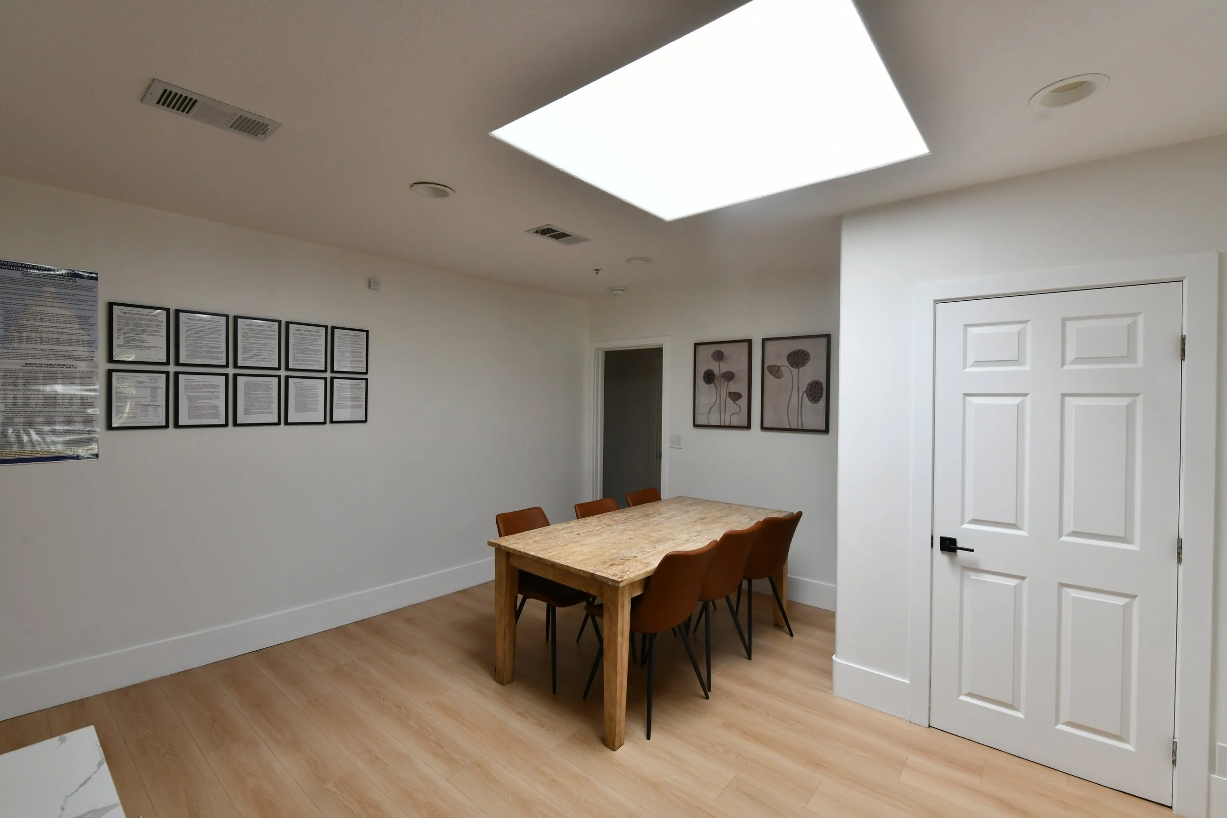 A room with white walls and a wooden floor, featuring a wooden table and six brown chairs. On the wall are framed pictures and a poster, with a large square ceiling light.
