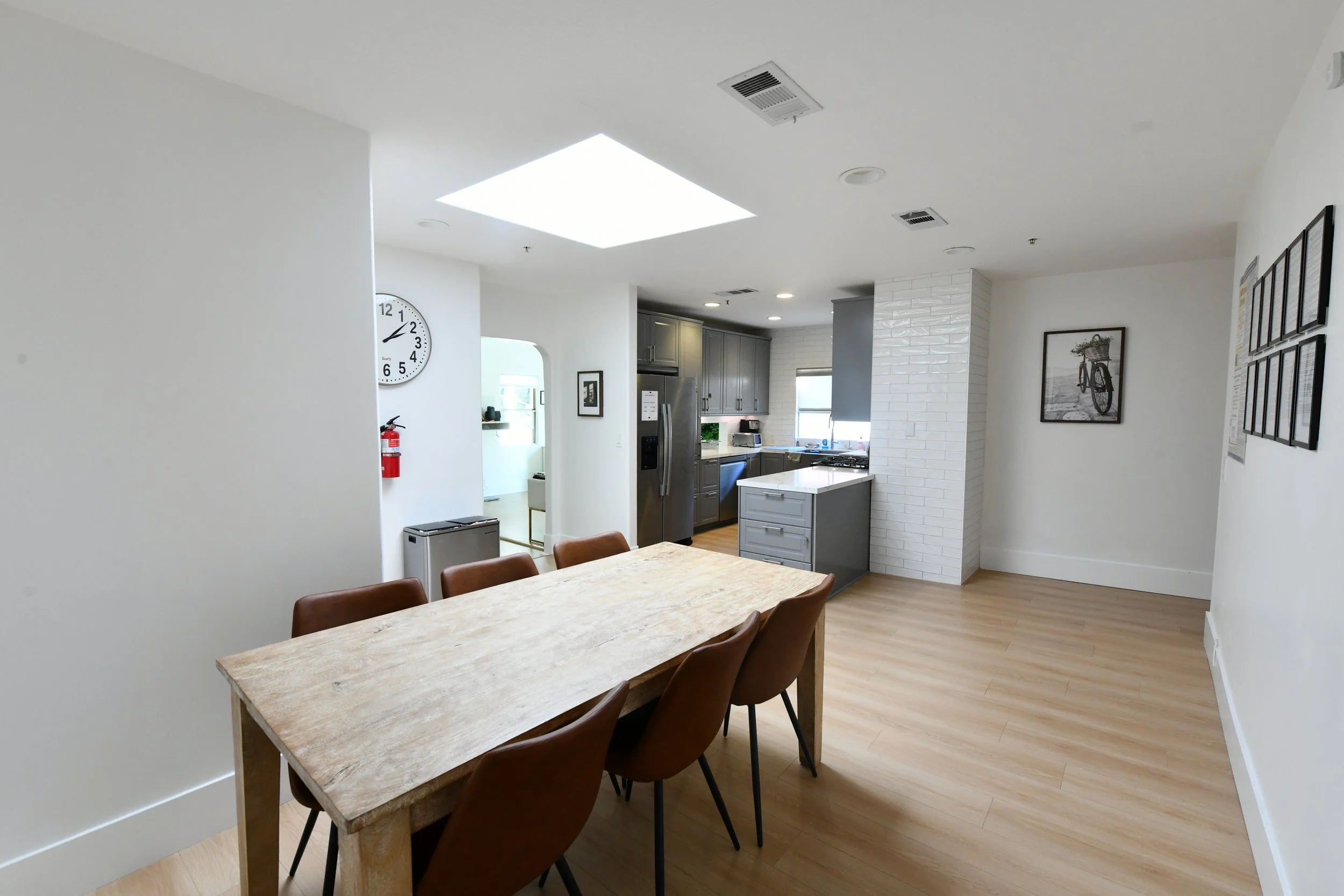 A bright, modern kitchen and dining area with a wooden table, six brown chairs, gray cabinets, a white brick wall, and a skylight in the ceiling.