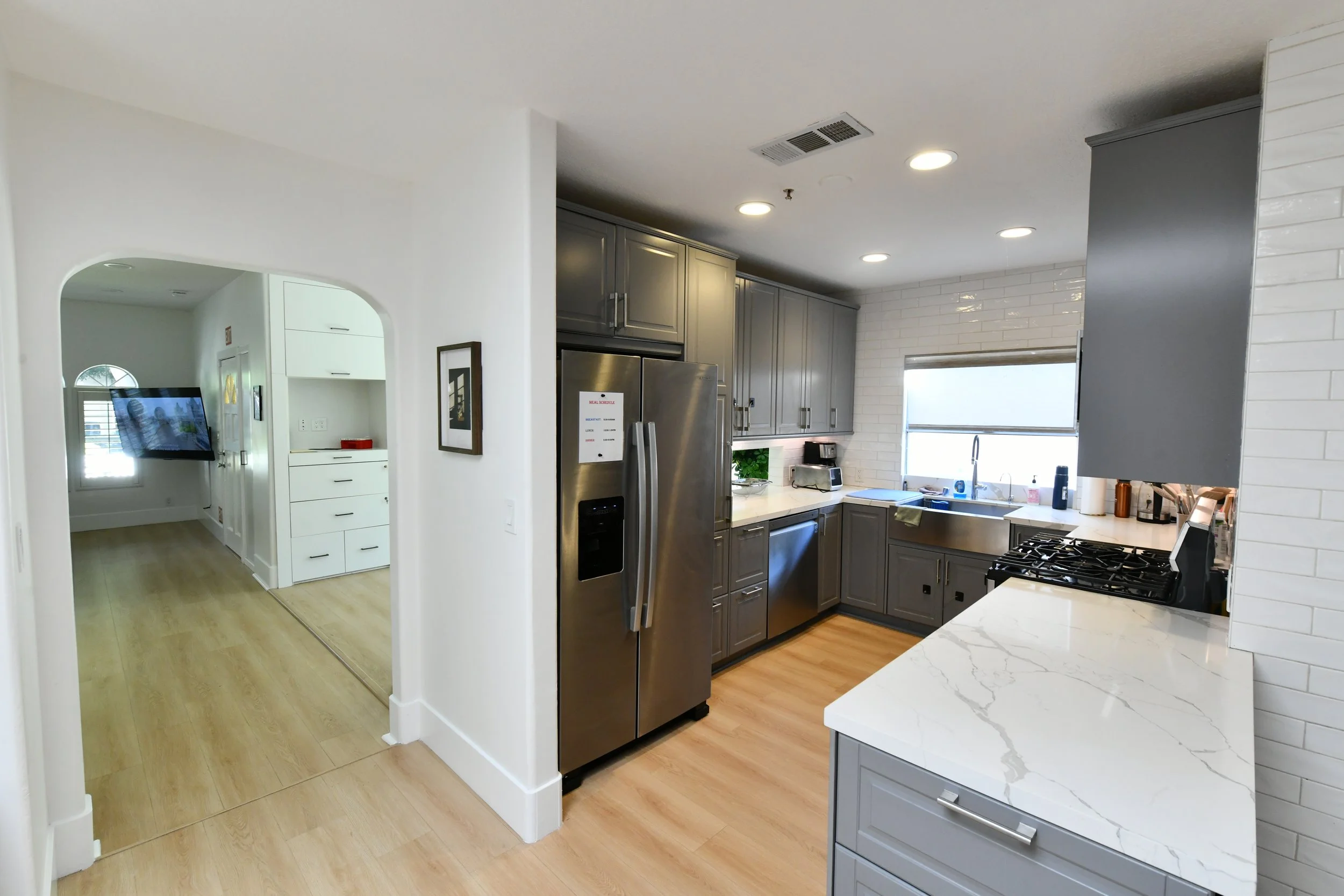 Modern kitchen with gray cabinets, stainless steel refrigerator, white marble countertop, subway tile backsplash, and a window above the sink.