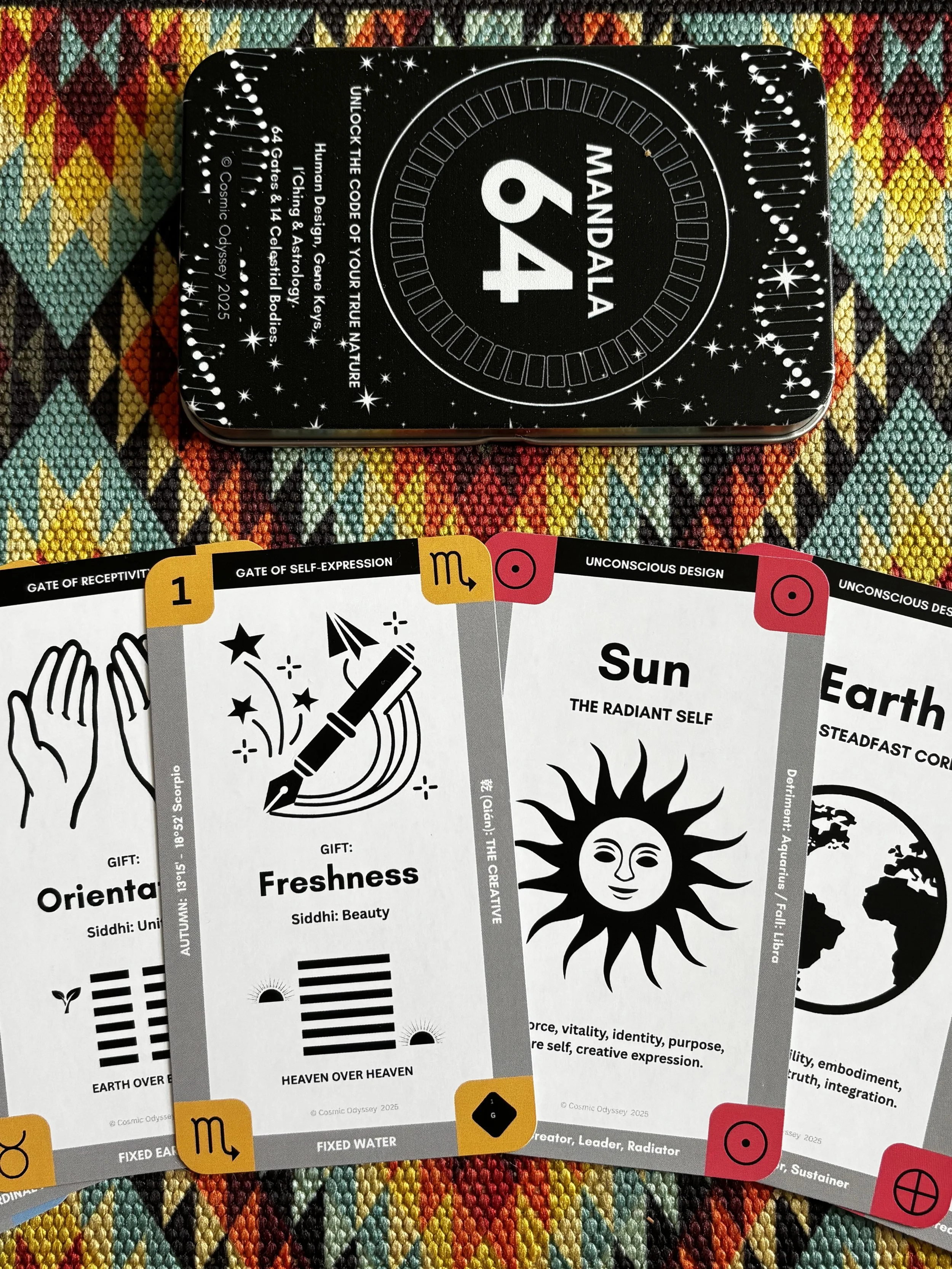 Cards from an oracle deck laid on a colorful woven cloth, with a metal tin box labeled "Mandala 64" in the background.